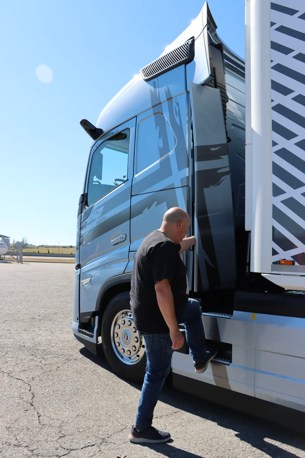Una persona se sube a un gran Volvo FH Aero estacionado en un día soleado, con un patrón de cuadros visible en el costado del vehículo. Probamos este modelo 460 I-Save para experimentar de primera mano su eficiencia y su elegante diseño.
