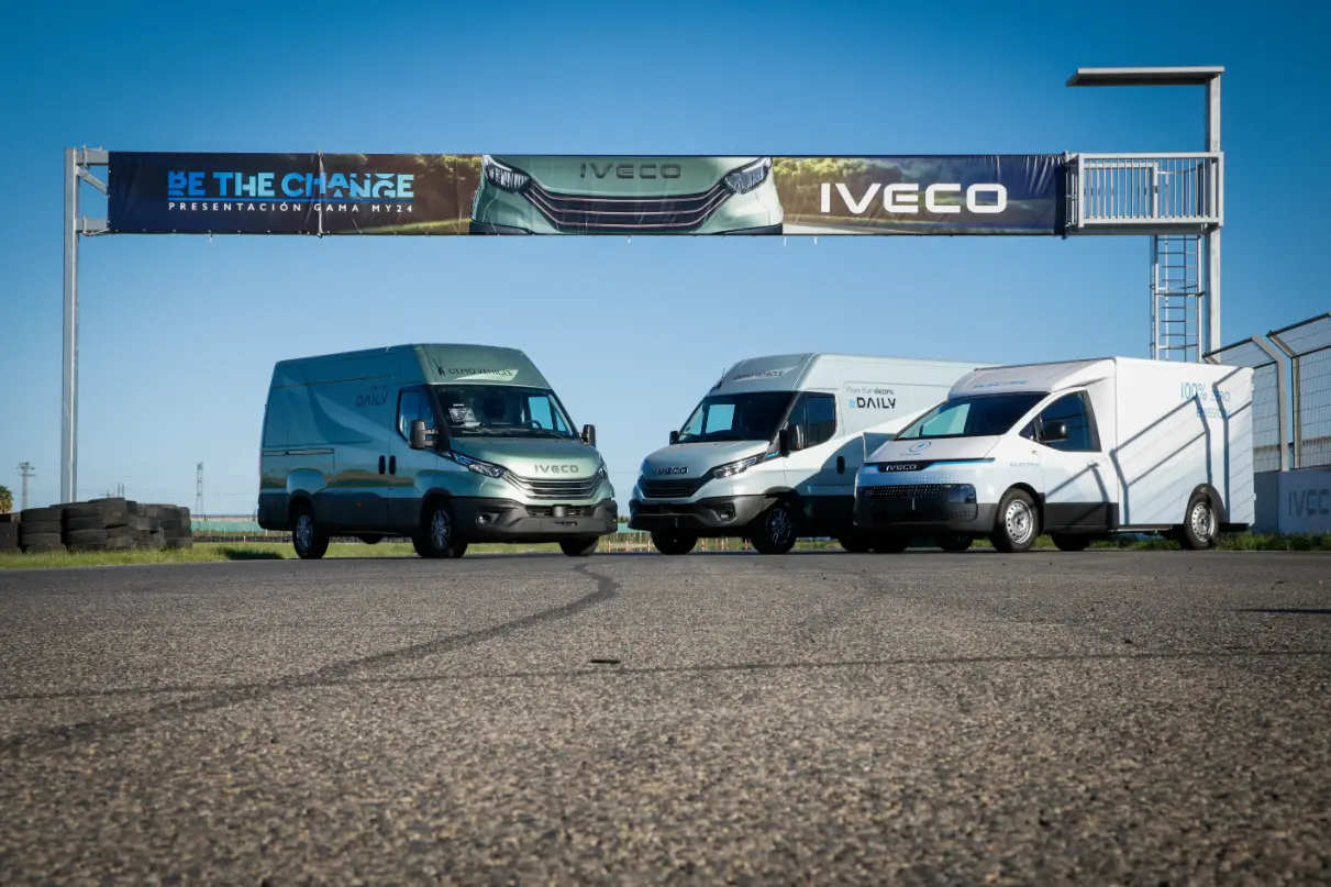 Tres furgonetas Iveco, de la nueva gama MY24, se encuentran aparcadas en una zona pavimentada bajo una pancarta en la que se puede leer "Be The Change". Al volante, su suavidad y eficiencia las hacen ideales tanto para trayectos urbanos como de larga distancia.