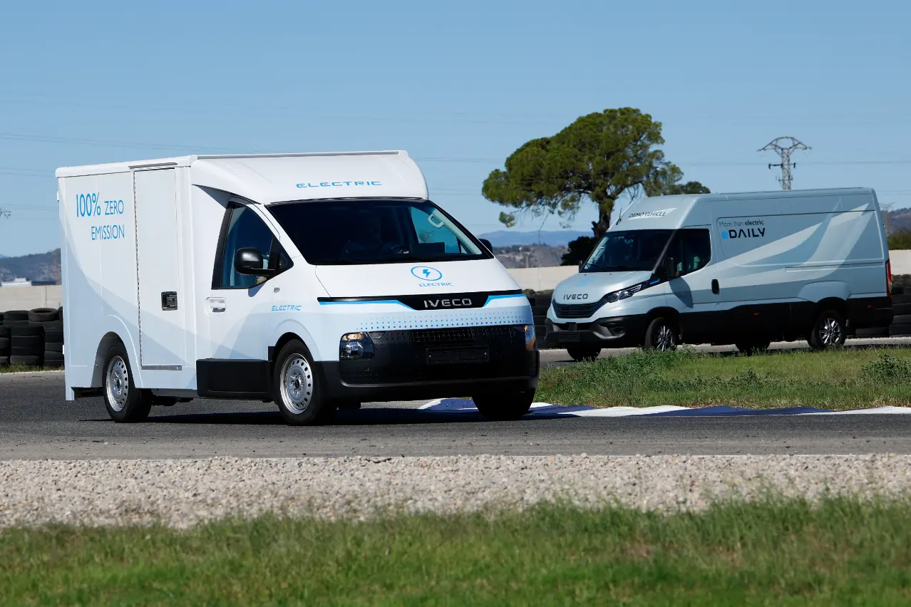 Dos furgonetas eléctricas Iveco de la gama MY24 adornan una escena de pista de carreras, una en movimiento y la otra estacionada, enmarcadas por un fondo de árboles y colinas onduladas.