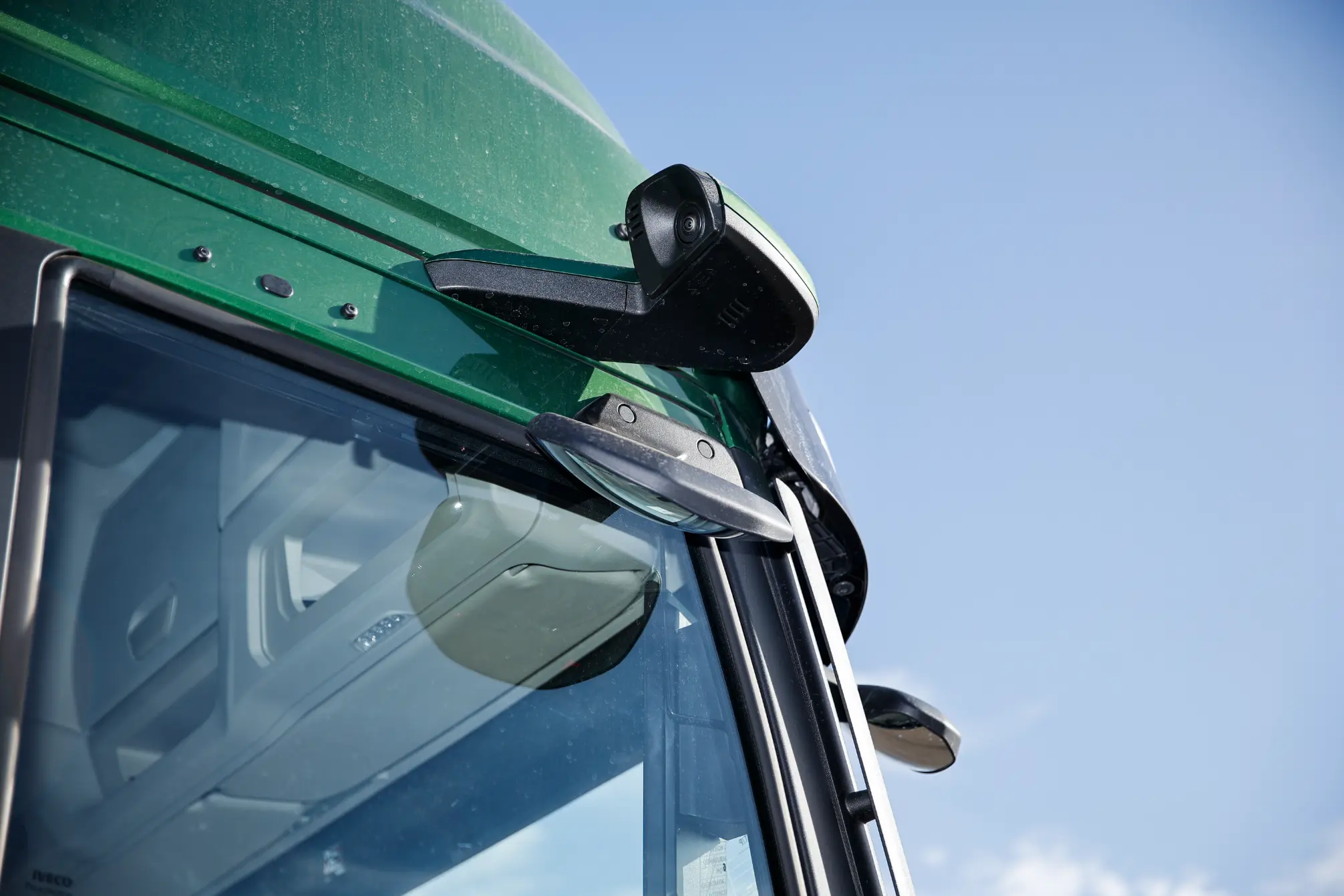 Primer plano de un autobús verde Iveco MY24 con cámaras montadas en el exterior, bajo un cielo azul claro.