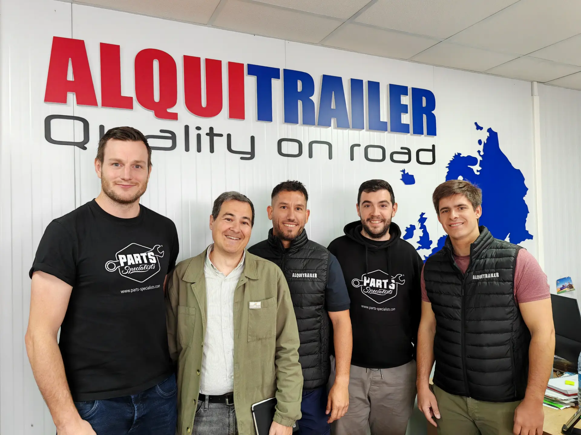 Cinco personas sonrientes se encuentran orgullosas frente a un cartel de "ALQUITRAILER" en el interior de un edificio, mostrando su experiencia con los productos Diesel Technic.