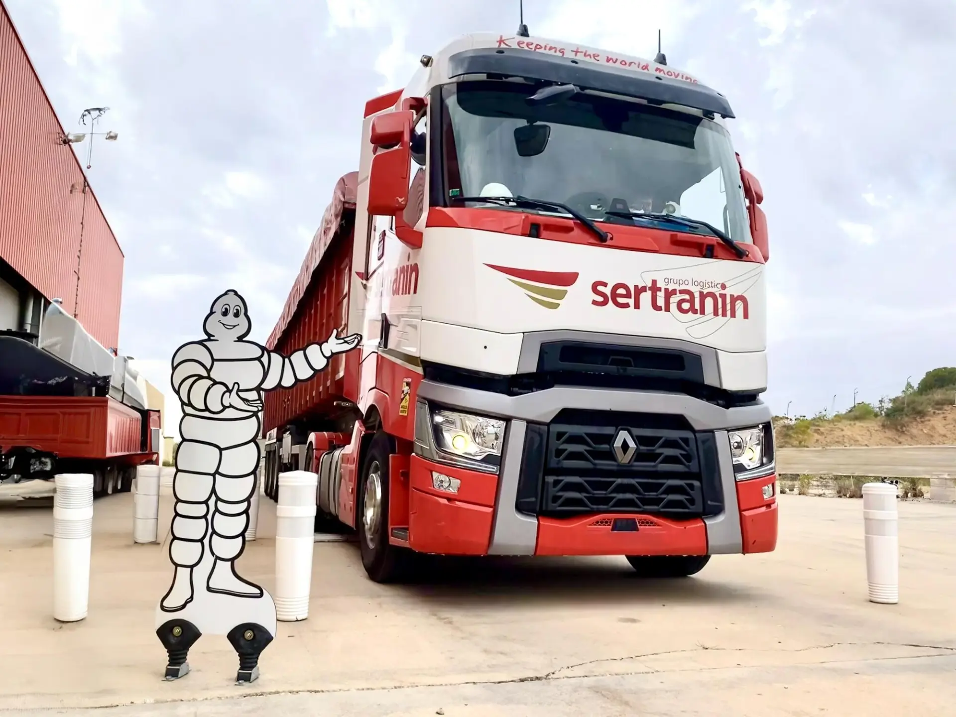 Un camión rojo está estacionado en el área de carga con una mascota de dibujos animados de neumáticos saludando alegremente, como si fuera un borrador automático que garantiza transiciones suaves.