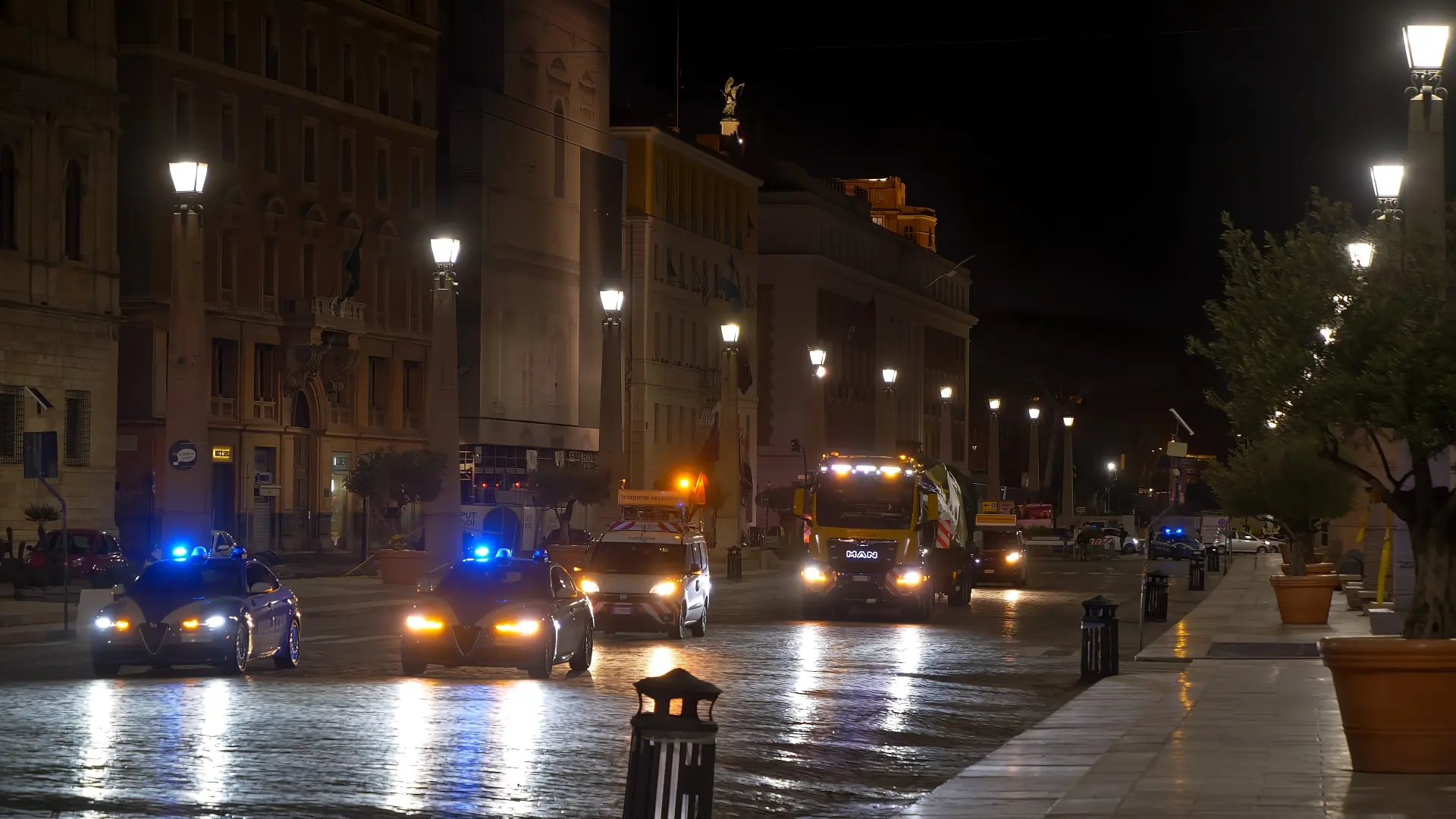 Calle de la ciudad de noche con coches de policía y un camión de basura, luces azules destellantes, reflejándose en el pavimento mojado como un borrador automático que corrige el caos.