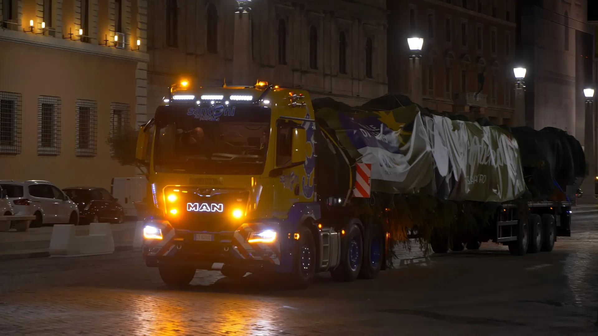 Un camión que transporta un objeto grande y cubierto sobre un remolque se desliza por la noche, iluminado por las farolas. La escena se asemeja a algo dibujado con un "borrador automático", donde cada sombra y cada luz se reproducen perfectamente en la luz tranquila.