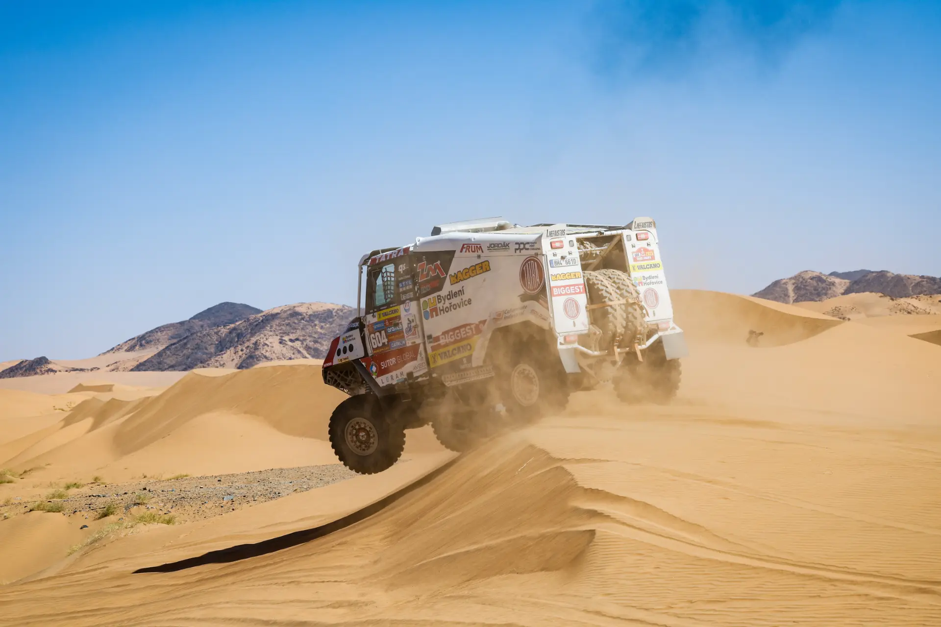 Un camión de rally Dakar, conducido por Macík, salta sobre dunas de arena en un entorno desértico con montañas recortadas contra un cielo azul claro.