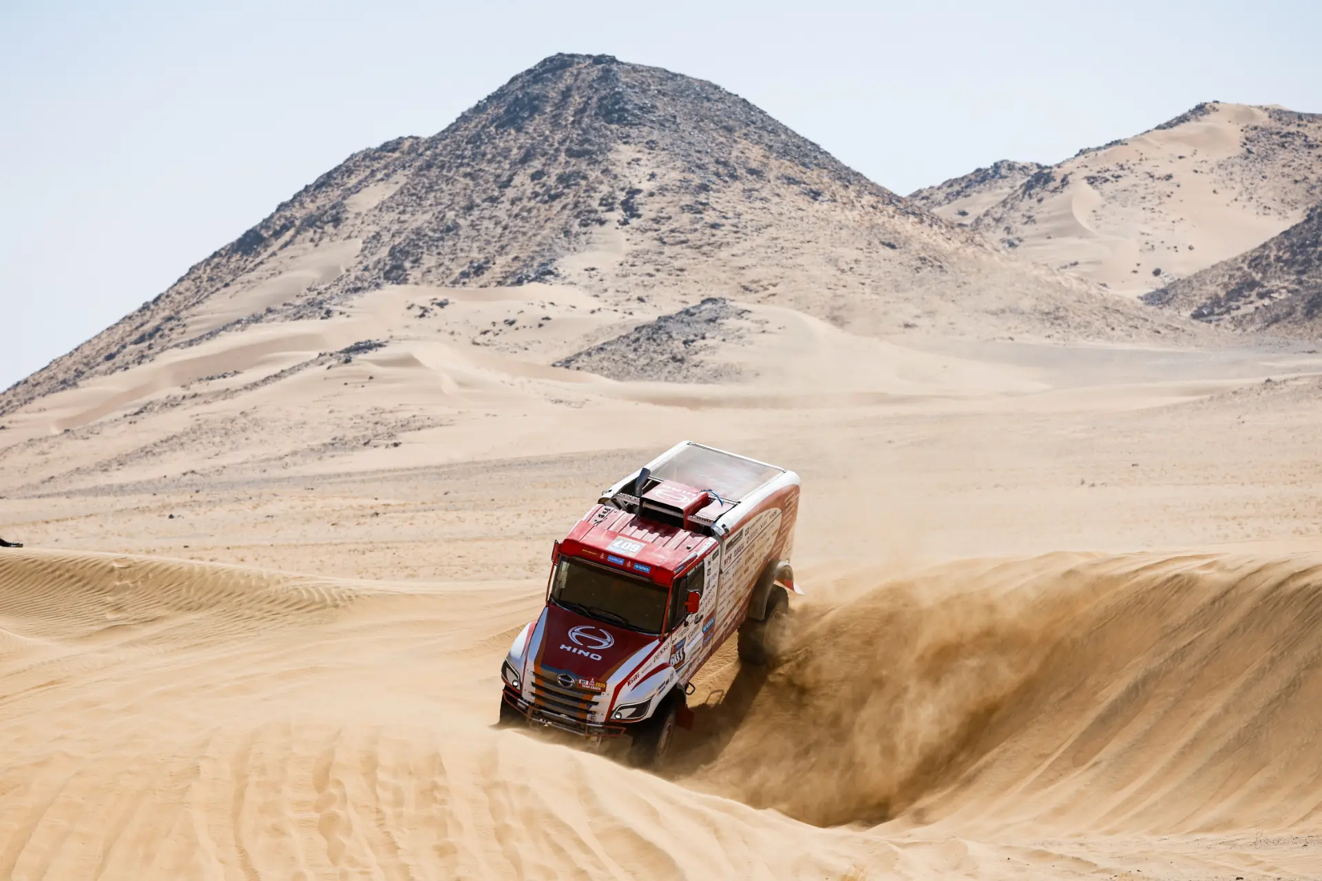 Un camión de rally rojo, que recuerda a los que condujo Macík en el Rally Dakar, levanta arena mientras avanza por las dunas del desierto con colinas rocosas de fondo.