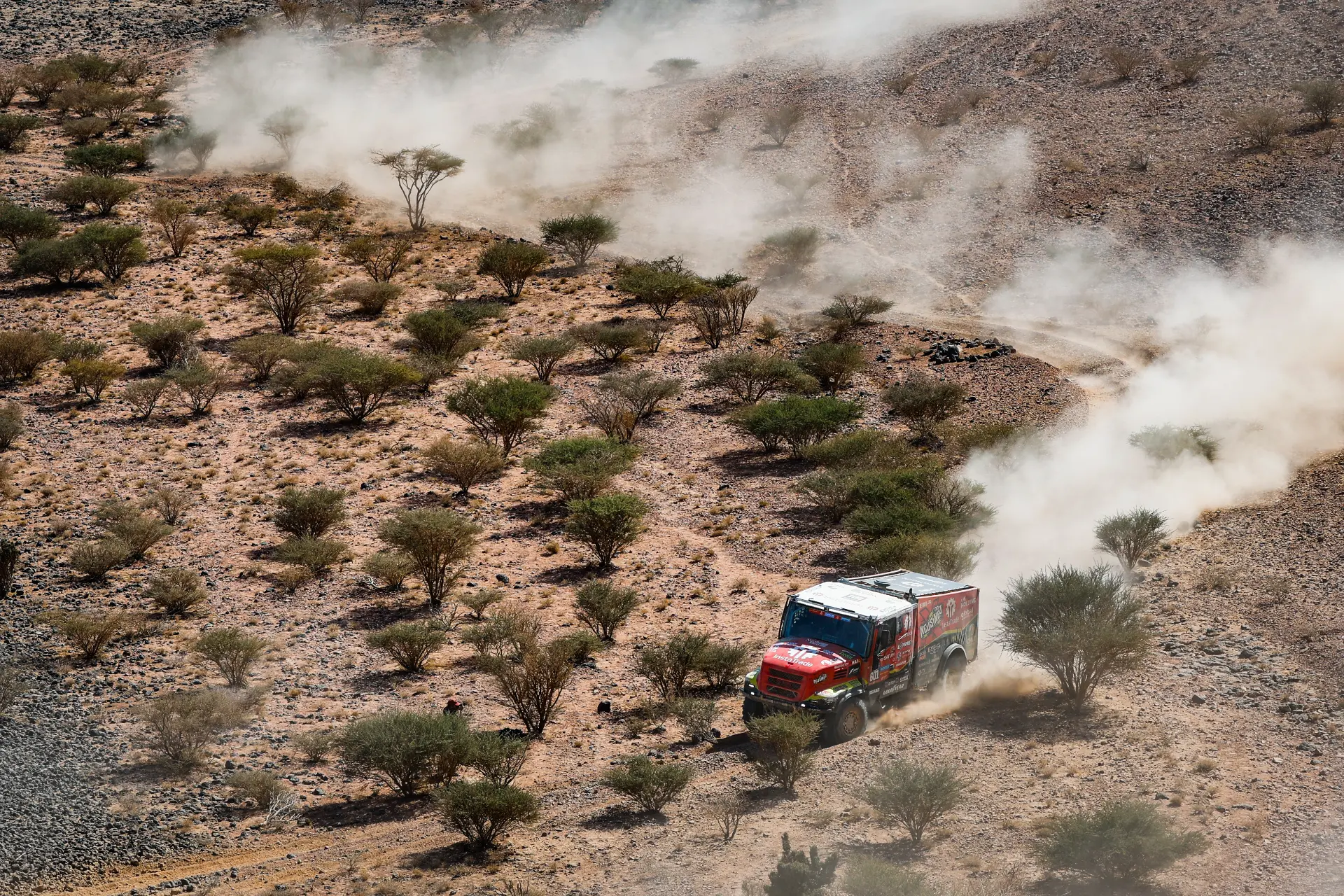 Un camión rojo levanta polvo como un participante del rally Dakar, conducido expertamente por Macík a través de un paisaje desértico con arbustos dispersos y terreno rocoso.