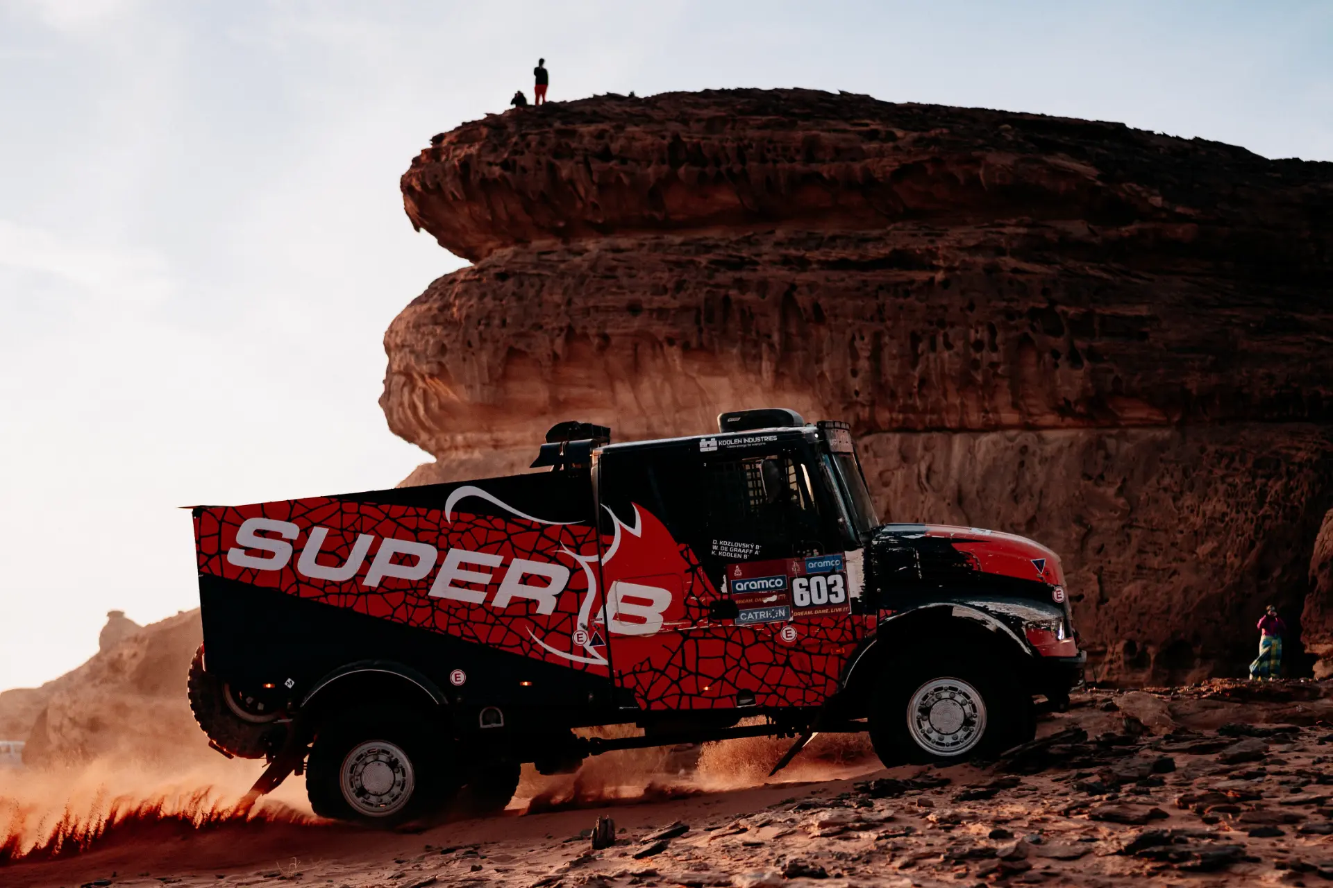 Un camión rojo y negro avanza rugiendo por el terreno accidentado y rocoso, transmitiendo la esencia del Rally Dakar con su feroz velocidad. Enormes formaciones rocosas se alzan como centinelas en el fondo, reflejando el espíritu intrépido de Macík mientras conquista los desafiantes obstáculos de la naturaleza.
