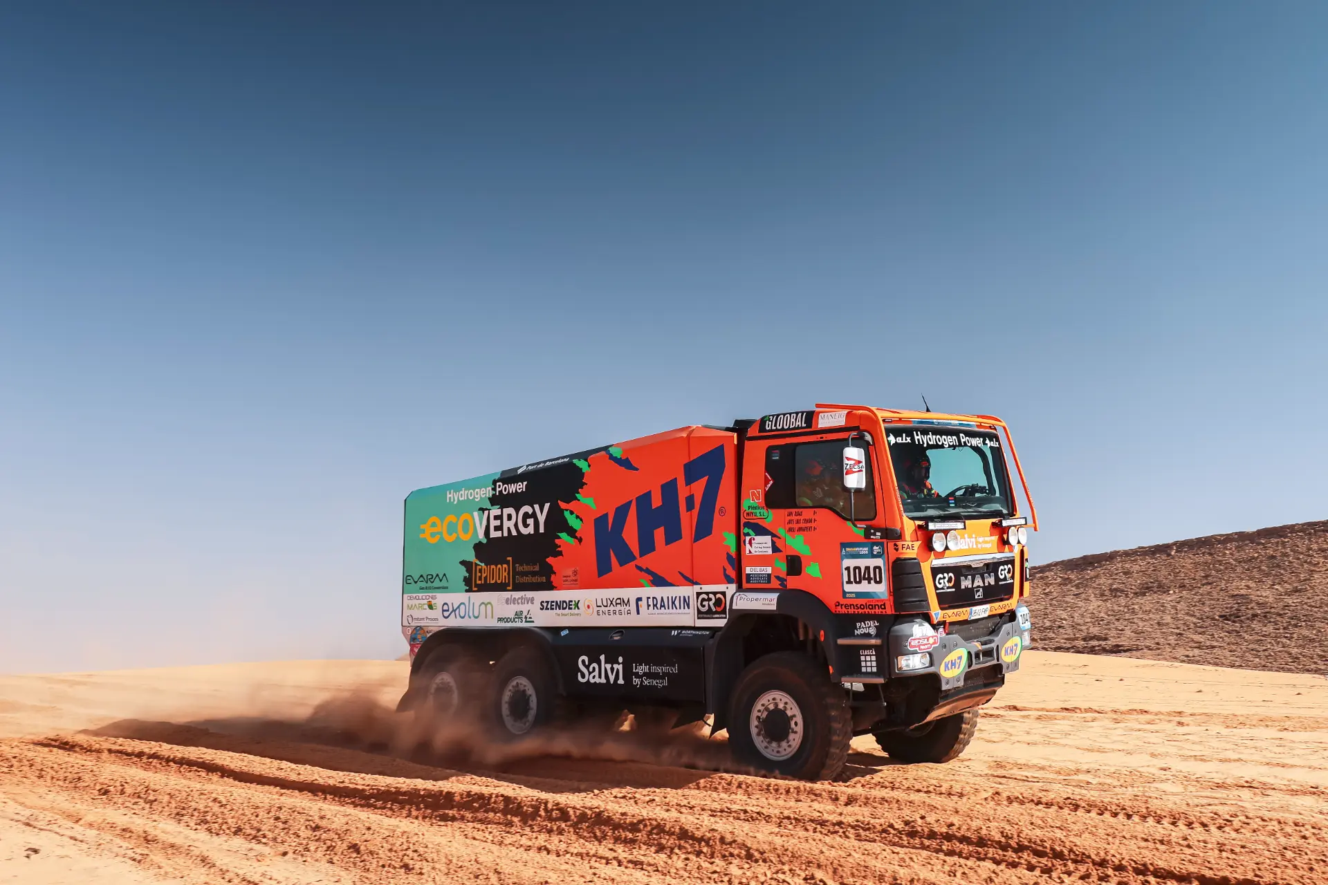 Un camión de carreras de colores brillantes, blasonado con los logotipos de Macík, avanza por el terreno desértico bajo un cielo azul claro, que recuerda a una emocionante etapa del rally Dakar.