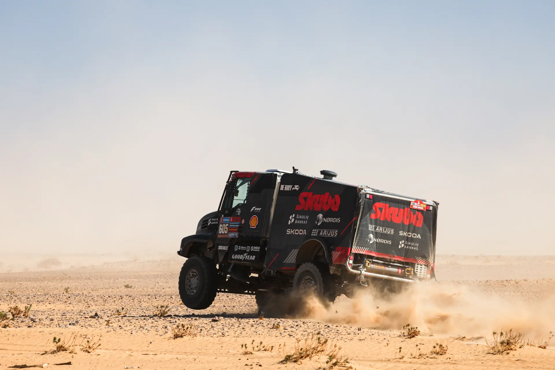 Un camión todoterreno con los logotipos de los patrocinadores y conducido por Macík avanza a toda velocidad por el terreno desértico del Rally Dakar, levantando nubes de polvo. La escasa vegetación salpica el suelo arenoso a su paso.