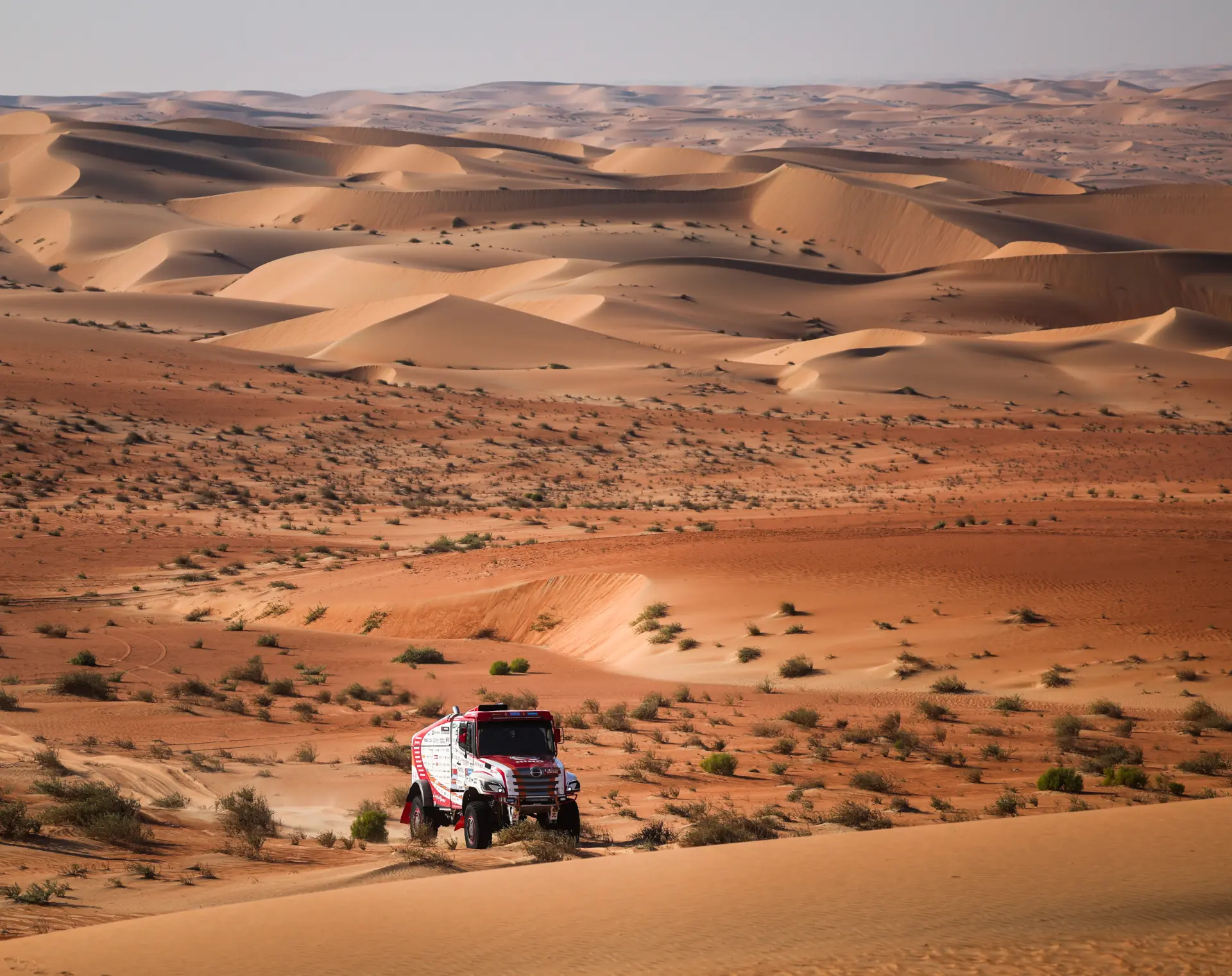 Un camión de rally conducido por Macík avanza a toda velocidad por vastas y onduladas dunas de arena bajo un cielo despejado en el paisaje del desierto, capturando el espíritu del Rally Dakar con la esperanza de un doblete triunfante.