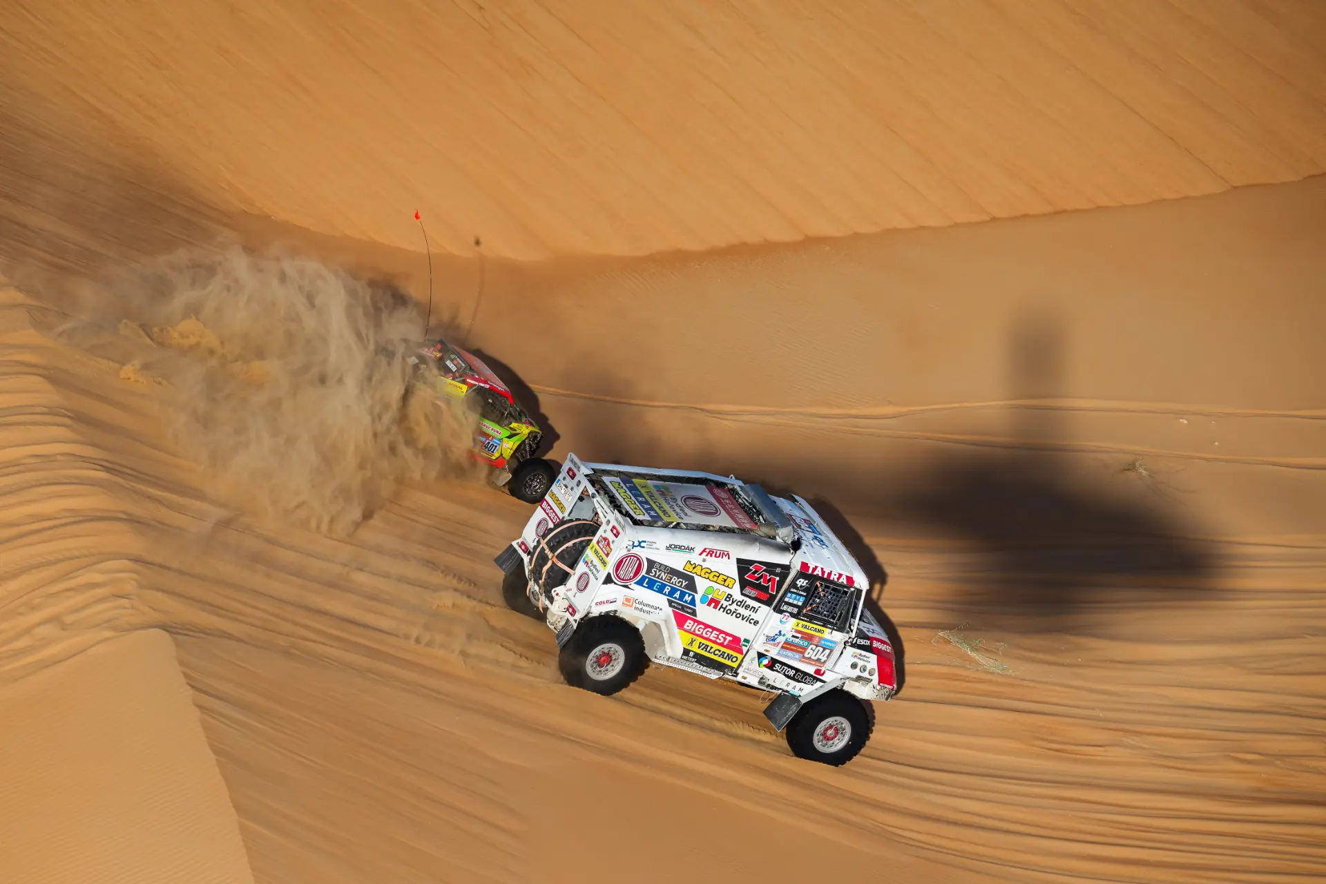 Dos camiones de rally, con nombres como Macík grabados en la historia de las carreras, retumban por las dunas del desierto del Rally Dakar. Levantan columnas de arena mientras la sombra de un helicóptero revolotea por el paisaje dorado: un verdadero espectáculo de Doblete.