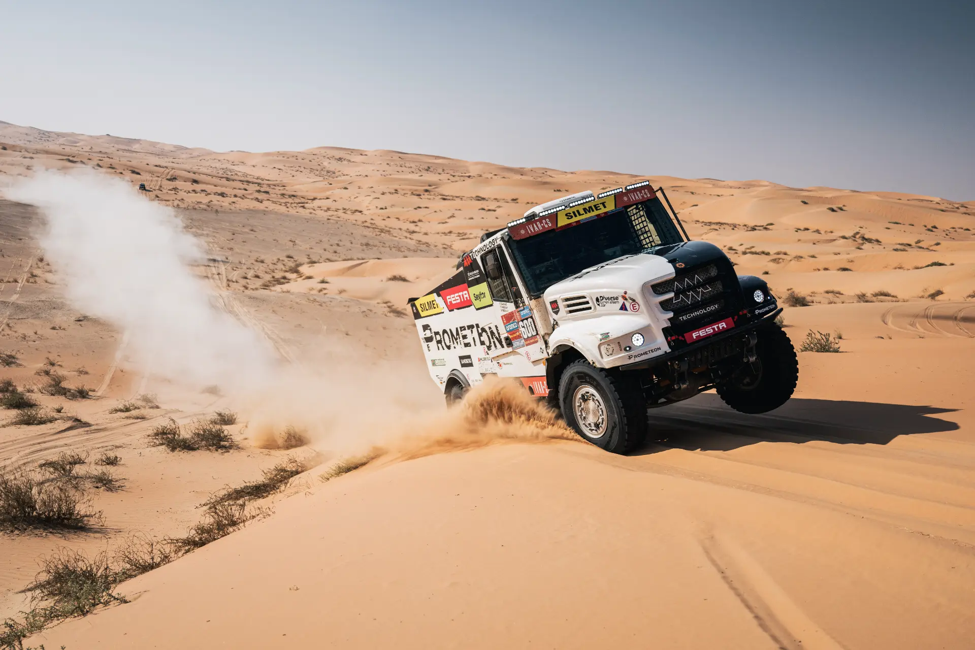 Camión todoterreno levantando arena mientras corre por el paisaje desértico bajo un cielo azul claro, que recuerda a un emocionante rally Dakar.