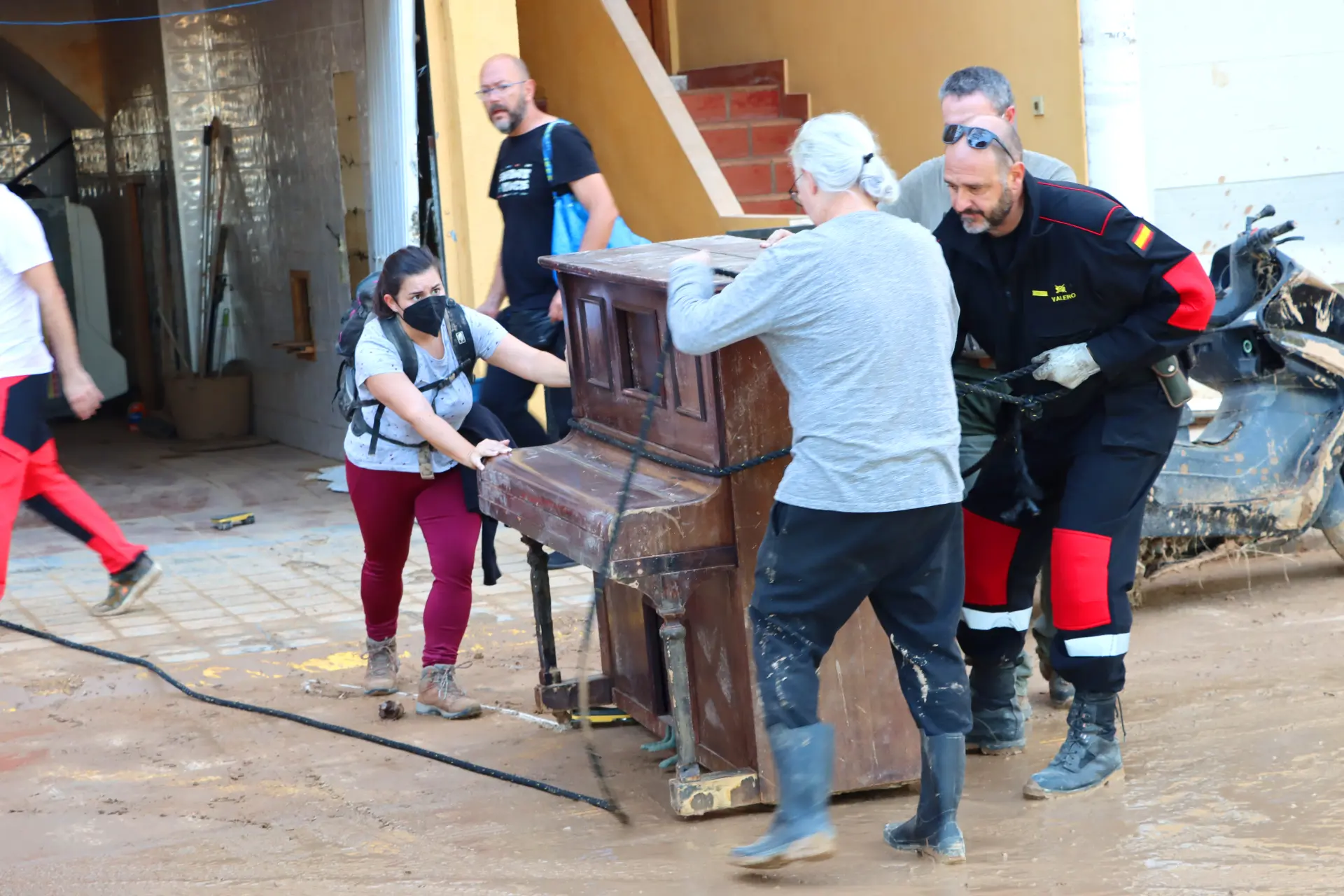 En una zona afectada por las inundaciones, personas del sector del transporte trabajan en equipo para mover un piano lleno de barro, con escombros esparcidos en el fondo.