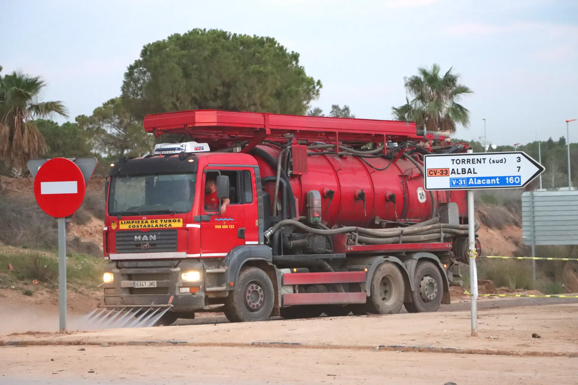 Un camión rojo de limpieza de alcantarillas circula por un camino de tierra bordeado de señales direccionales y palmeras ondulantes, representando el trabajo esencial dentro del sector del transporte.