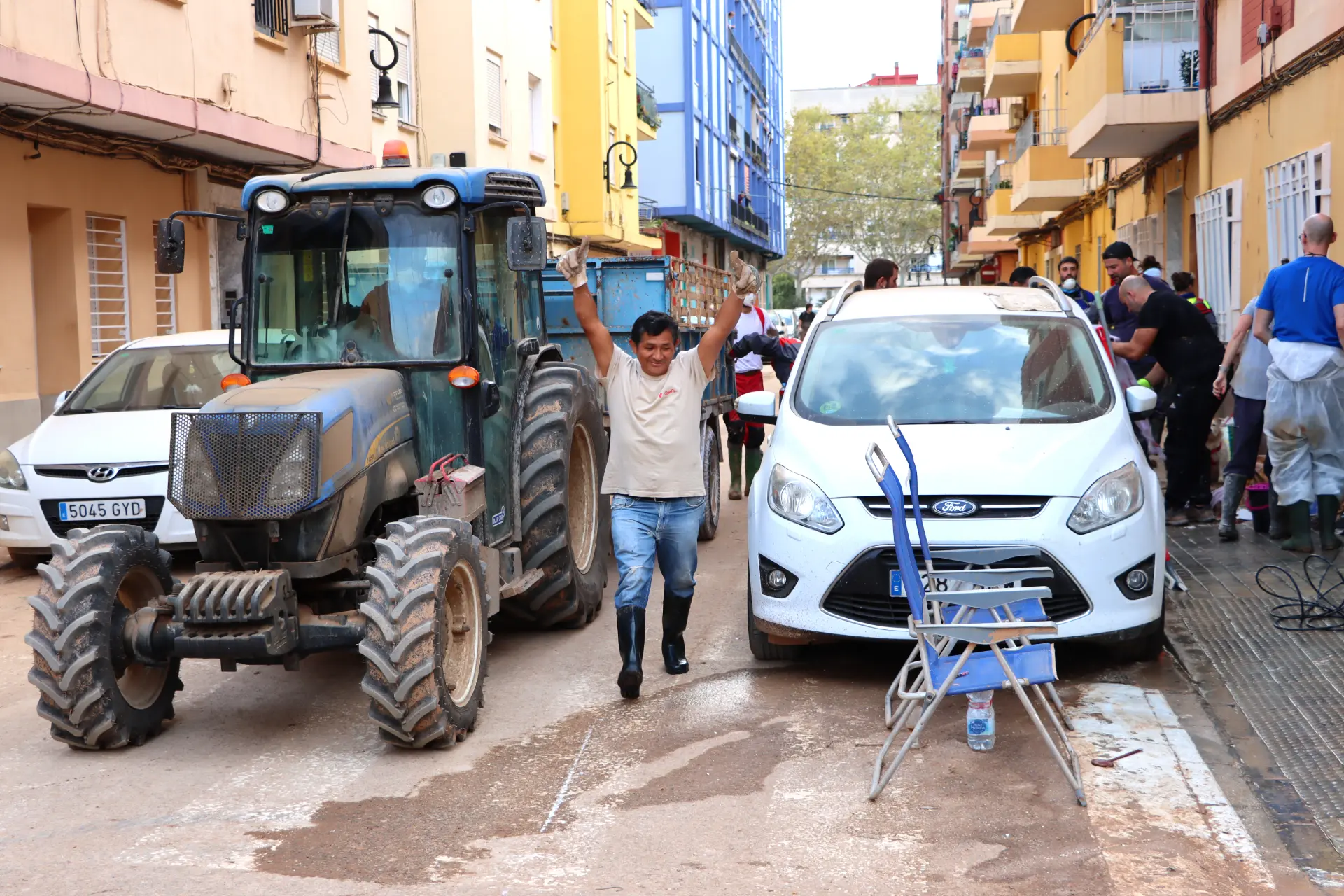 Un hombre se regocija en medio de un tractor y un automóvil en una calle empapada por la lluvia, mientras la gente trabaja incansablemente para limpiar en el fondo, mostrando resiliencia ante la dana dentro del sector del transporte.