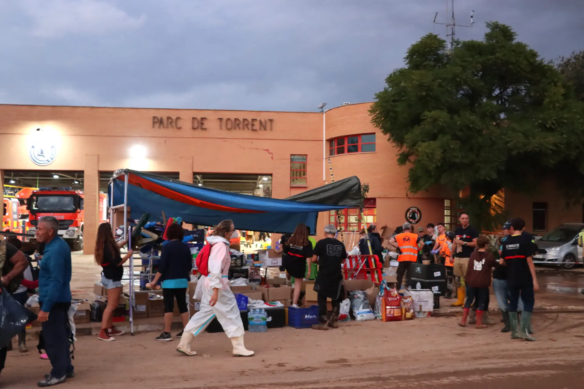 Los voluntarios distribuyen suministros en el exterior de un edificio marcado como "Parc de Torrent" mientras los vehículos de emergencia responden a las consecuencias de un reciente daño que afectó al sector del transporte.