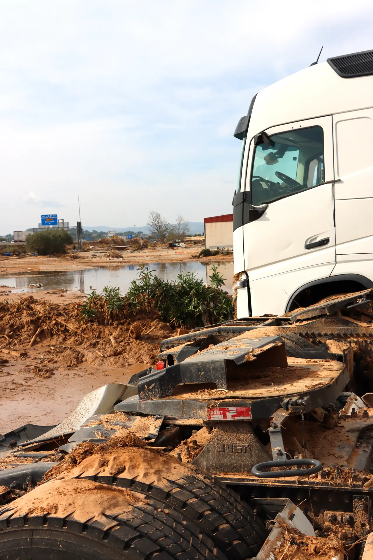 Un camión del sector del transporte se encuentra atascado en un terreno fangoso cerca del agua, con edificios y vegetación exuberante al fondo, una situación inesperada debido a las recientes condiciones de dana.