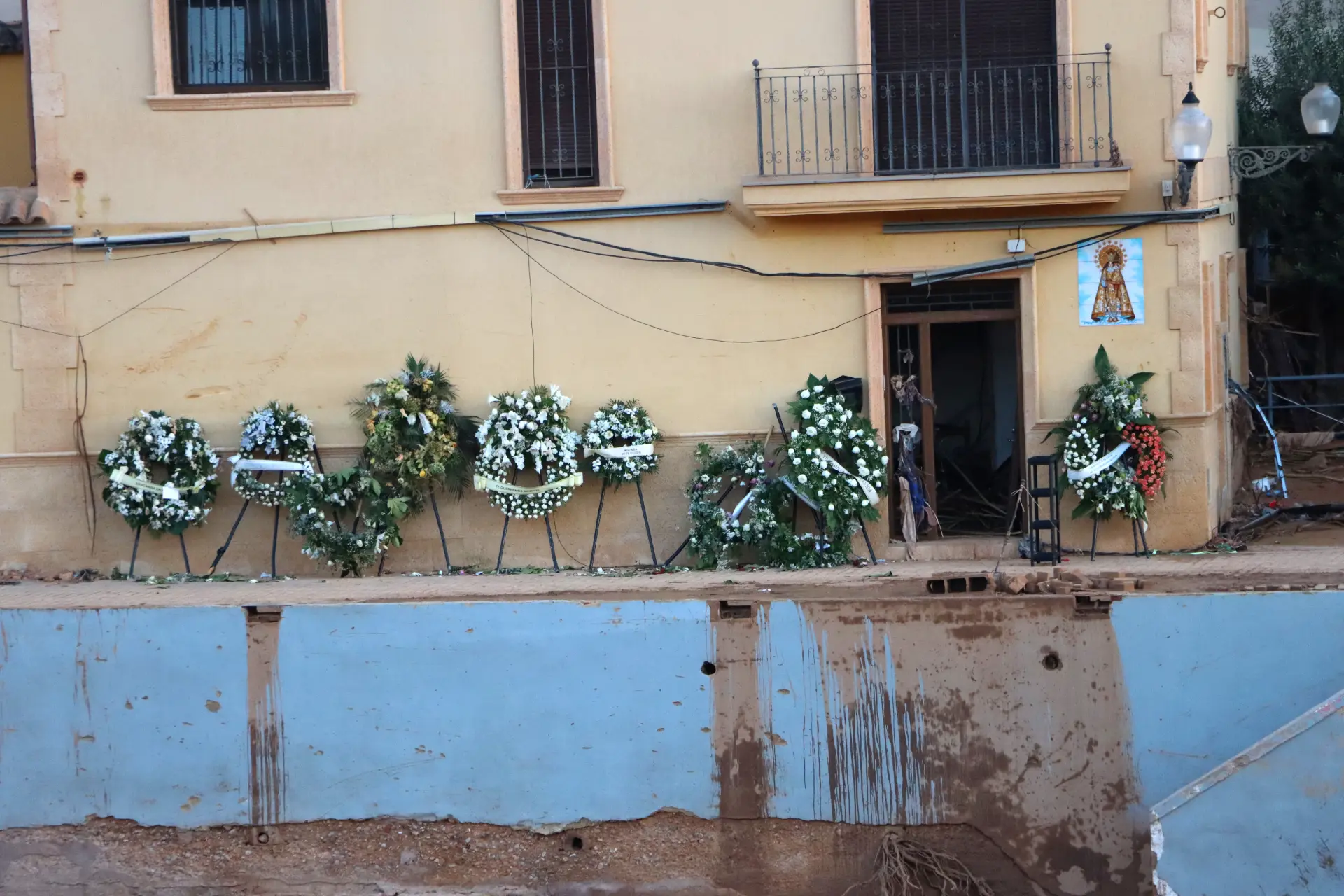 Coronas de flores se alinean en la pared de un edificio de color beige sobre un área dañada y fangosa con un revestimiento azul expuesto, que recuerda los caminos desgastados en el sector del transporte de Dana.