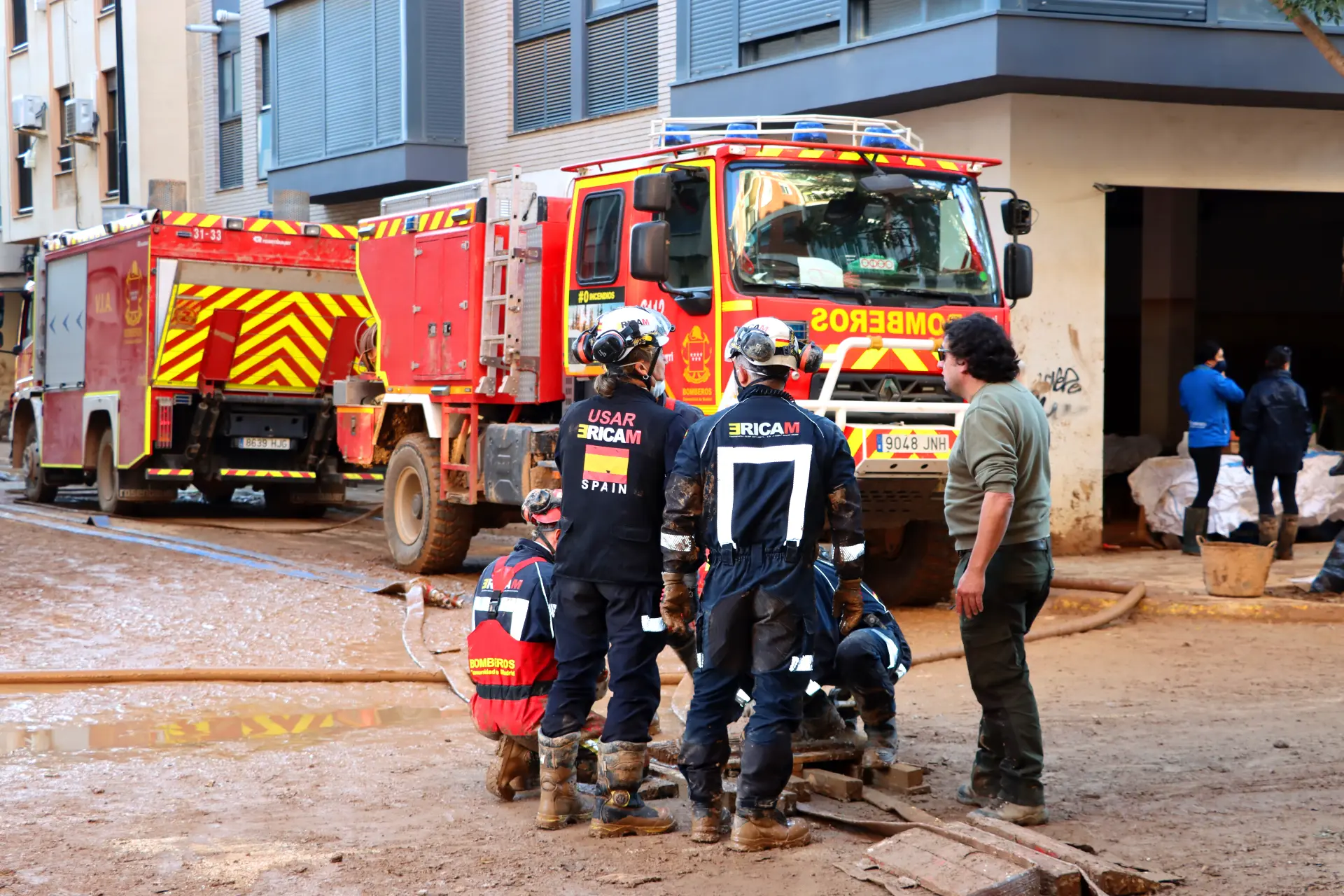 Los equipos de emergencia con equipo de protección están cerca de un camión de bomberos en una calle fangosa, posiblemente debido a los daños causados por Dana; una persona está agachada.