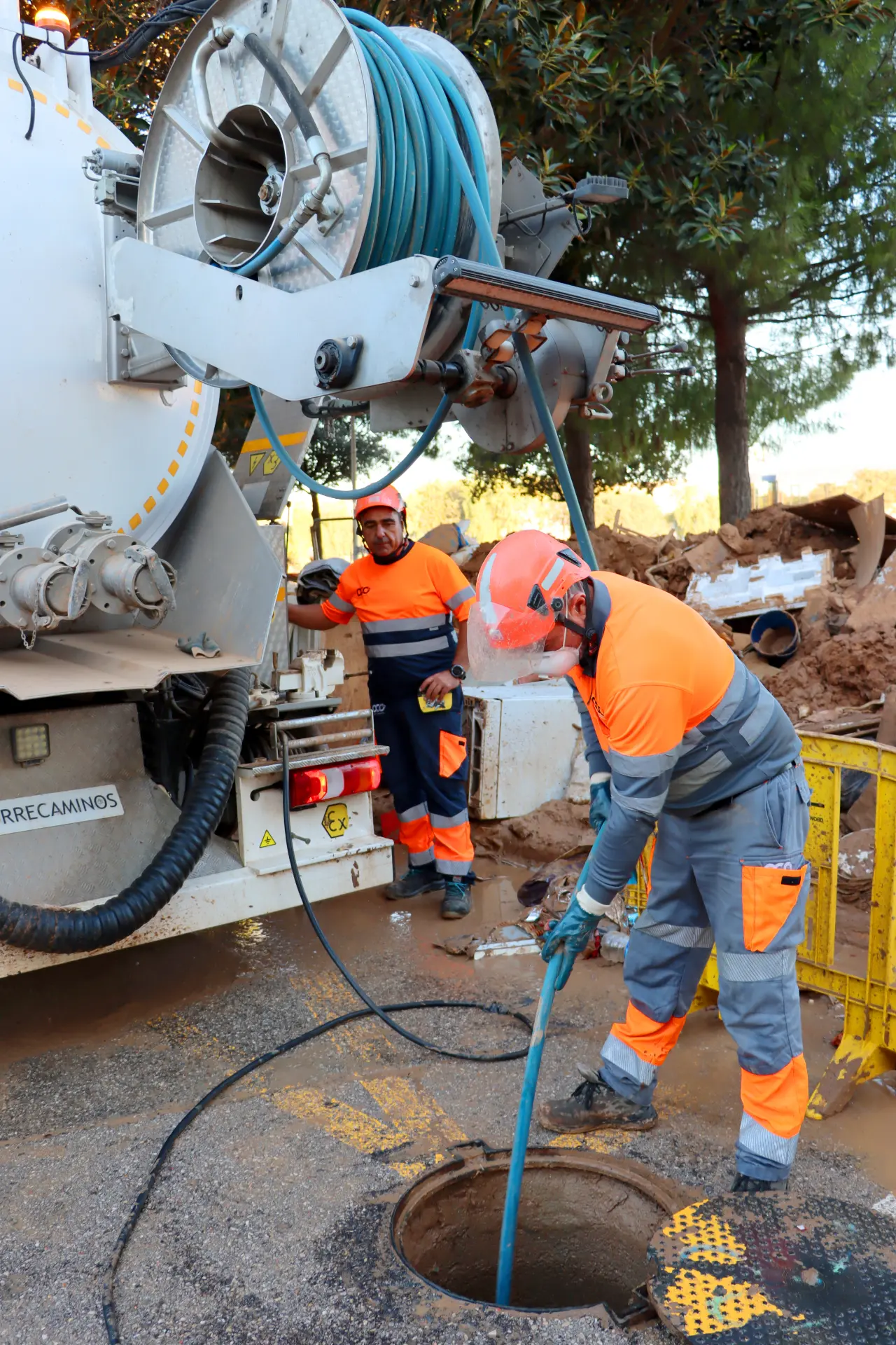 Dos trabajadores con uniformes de color naranja utilizan equipo para limpiar un pozo en una calle, con un gran vehículo del sector de transporte cerca, asegurándose de que la infraestructura permanezca intacta.