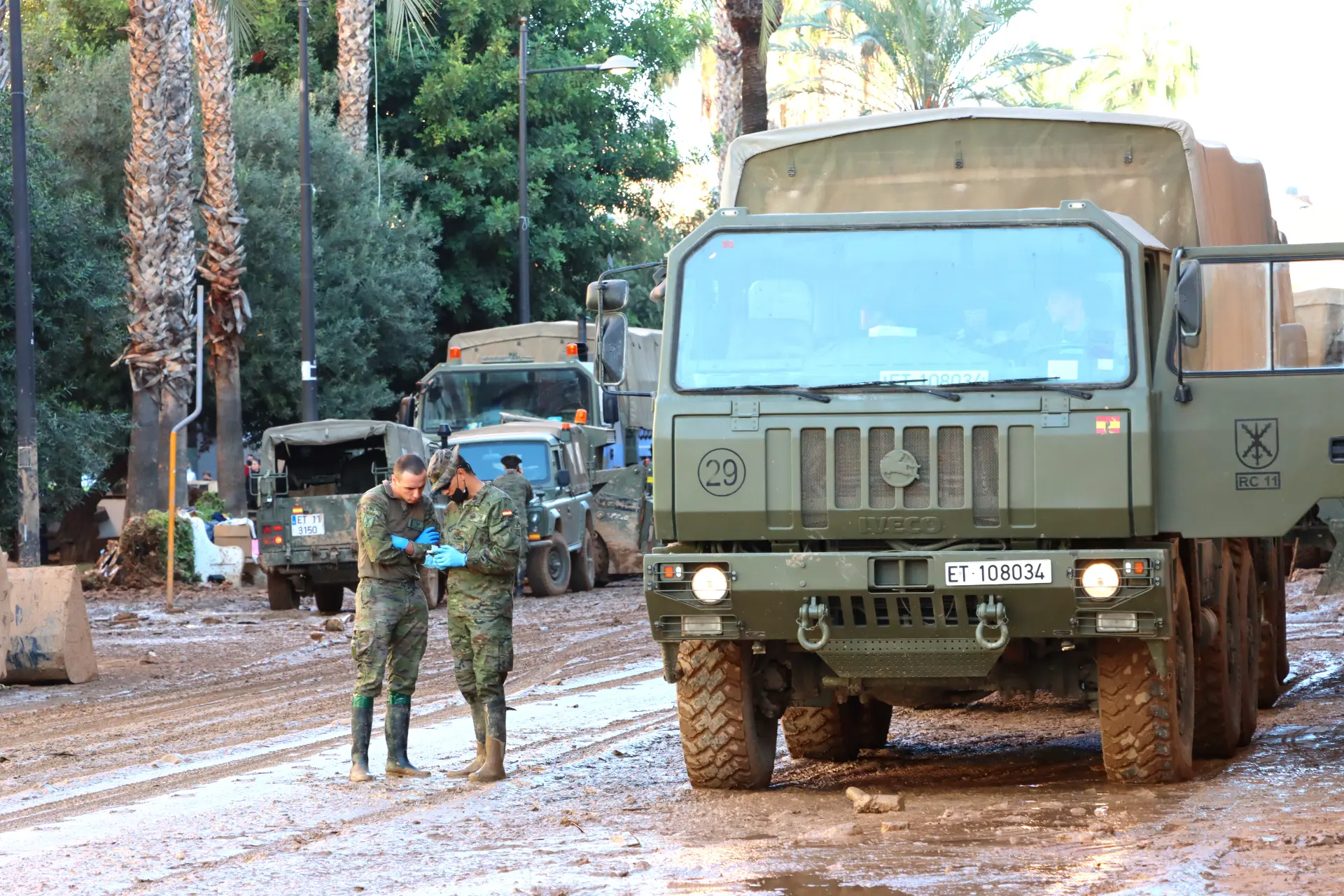 Soldados con uniformes embarrados se encuentran junto a un camión militar en un camino de tierra, rodeados de árboles y más vehículos, encarnando la resiliencia del sector del transporte en terrenos desafiantes.