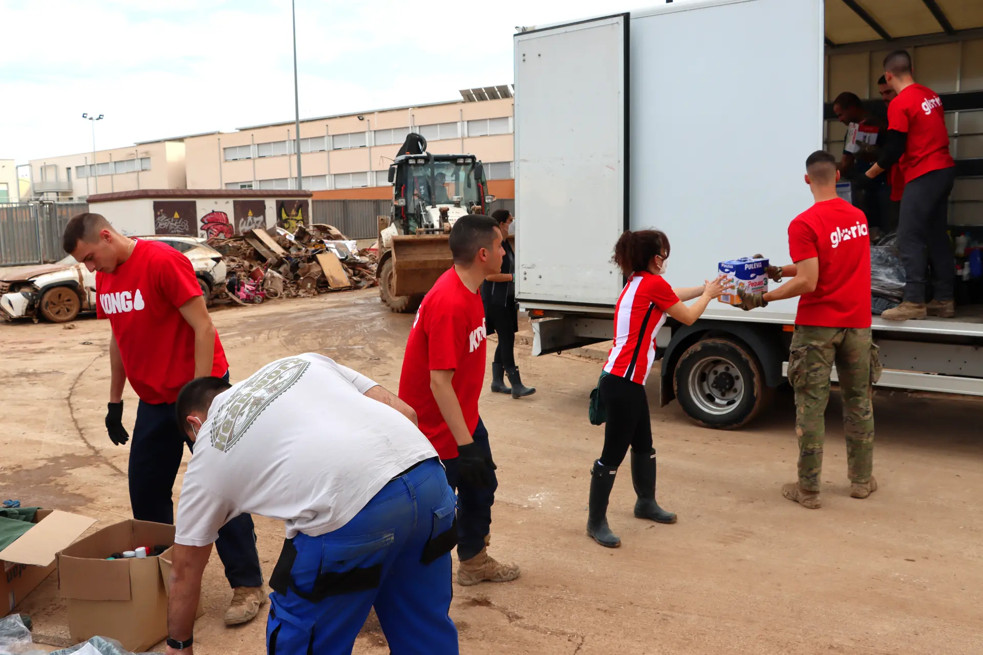 Voluntarios con camisas rojas, parte del equipo dana, están descargando suministros de un camión cerca de una excavadora y escombros, apoyando al sector del transporte con sus eficientes esfuerzos.
