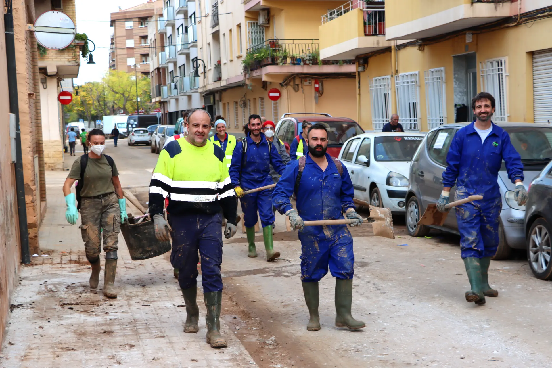 Un grupo de personas con ropa de trabajo y botas avanza por una calle embarrada, cargando palas y baldes. Detrás de ellos, los edificios se yerguen mientras avanzan en su tarea en medio de los desafíos que trajo consigo el reciente dana.