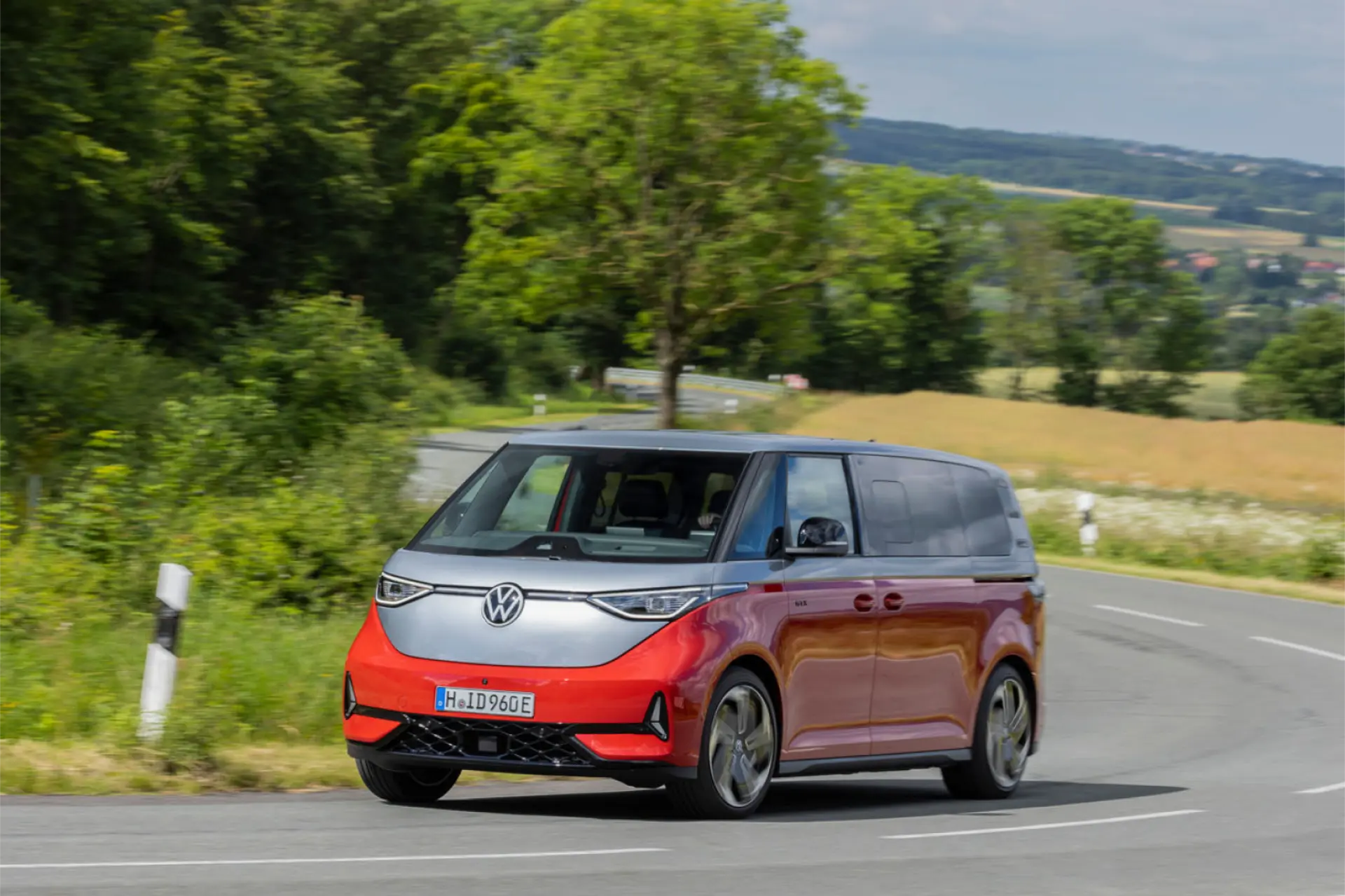 Una llamativa furgoneta Volkswagen roja, famosa por su tracción total, circula con suavidad por una carretera rural flanqueada por una exuberante vegetación y onduladas colinas de fondo.