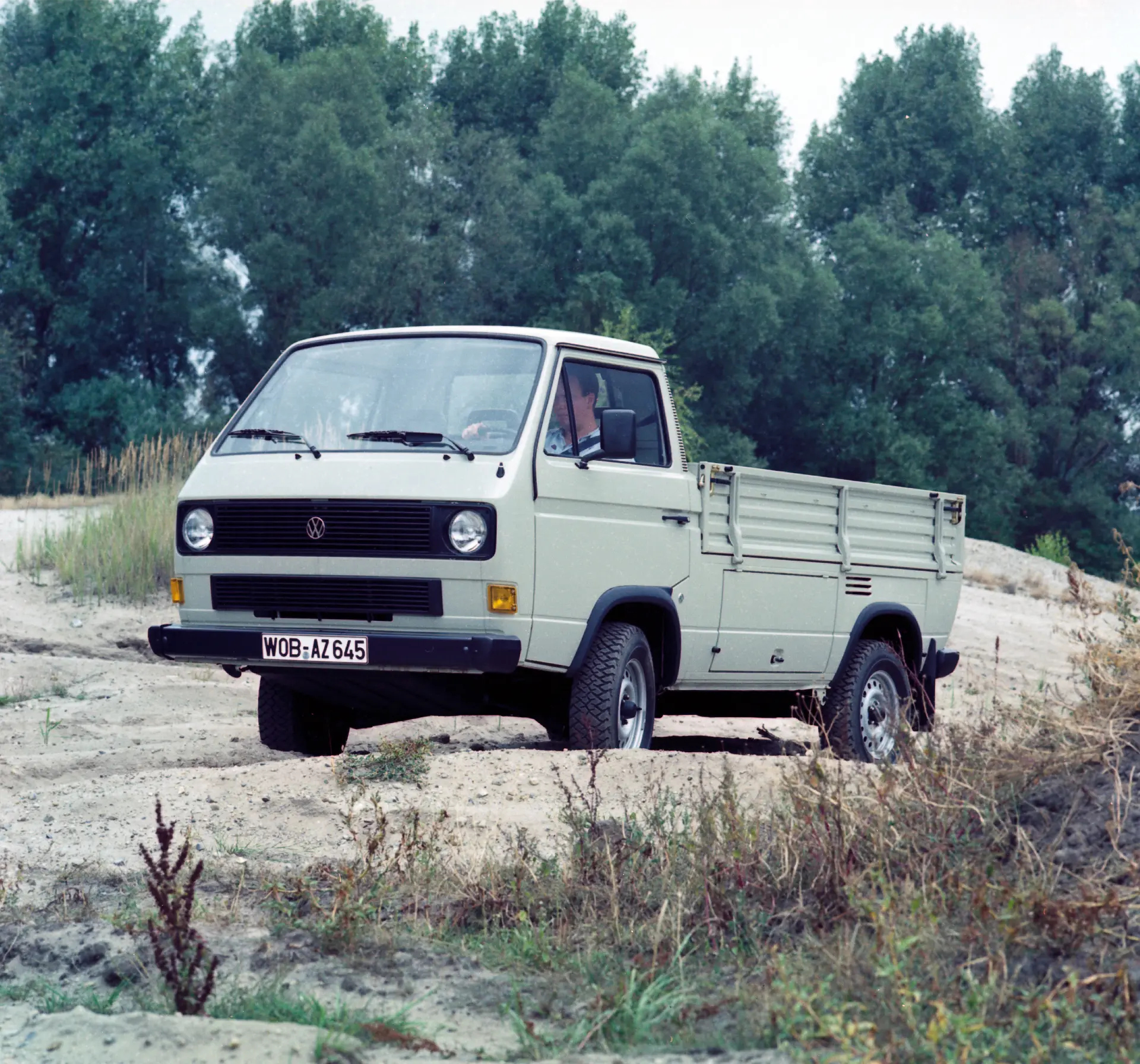 Una camioneta utilitaria Volkswagen clásica, con tracción total, está estacionada en un terreno arenoso con árboles al fondo, mostrando su legado en Vehículos Comerciales.