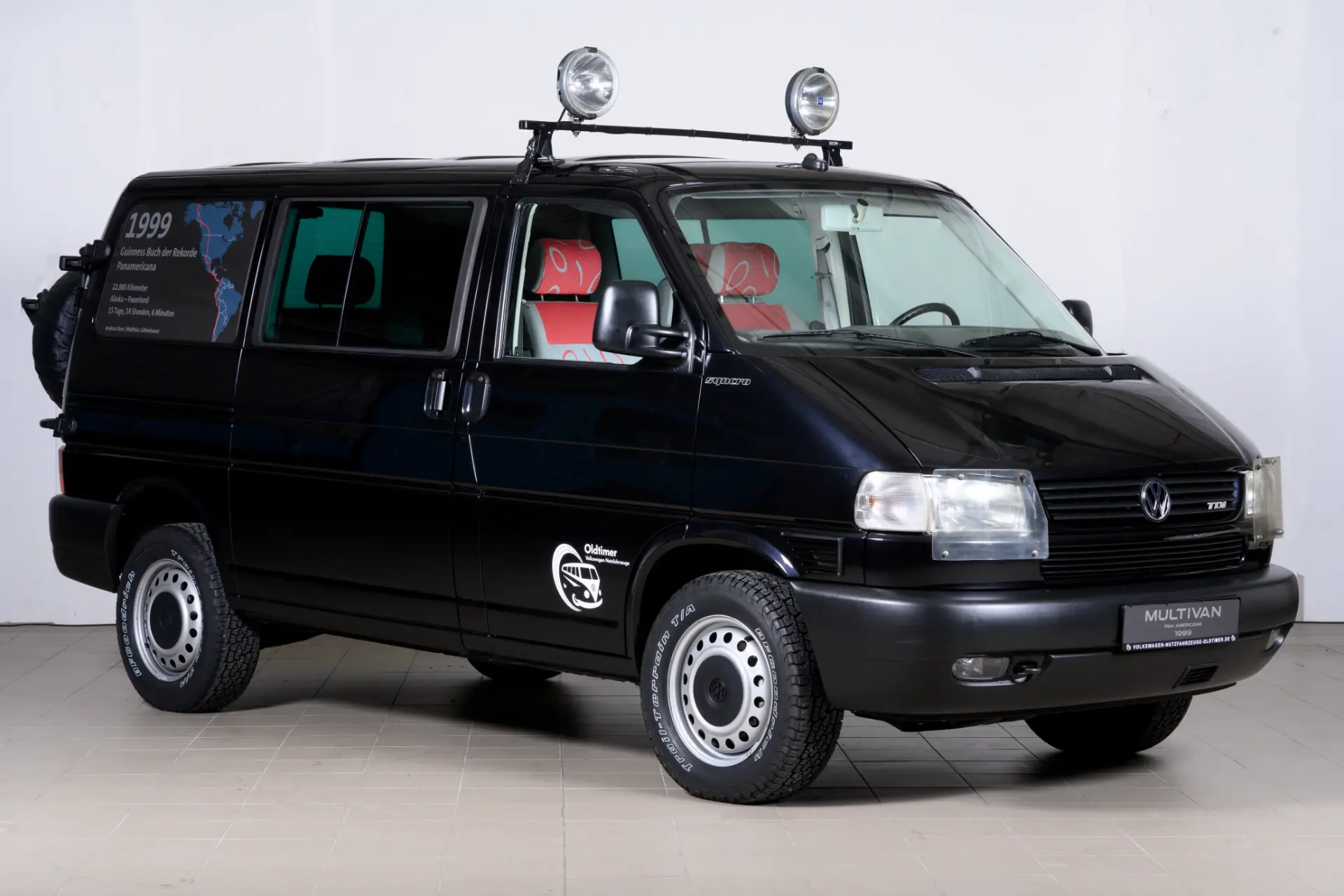 La Volkswagen T4 Multivan negra, con luces en el techo y un adhesivo lateral, se exhibe con elegancia en la sala de exposición. Este vehículo versátil pertenece a la línea Vehículos Comerciales y ofrece tracción total opcional para un mejor rendimiento de conducción.