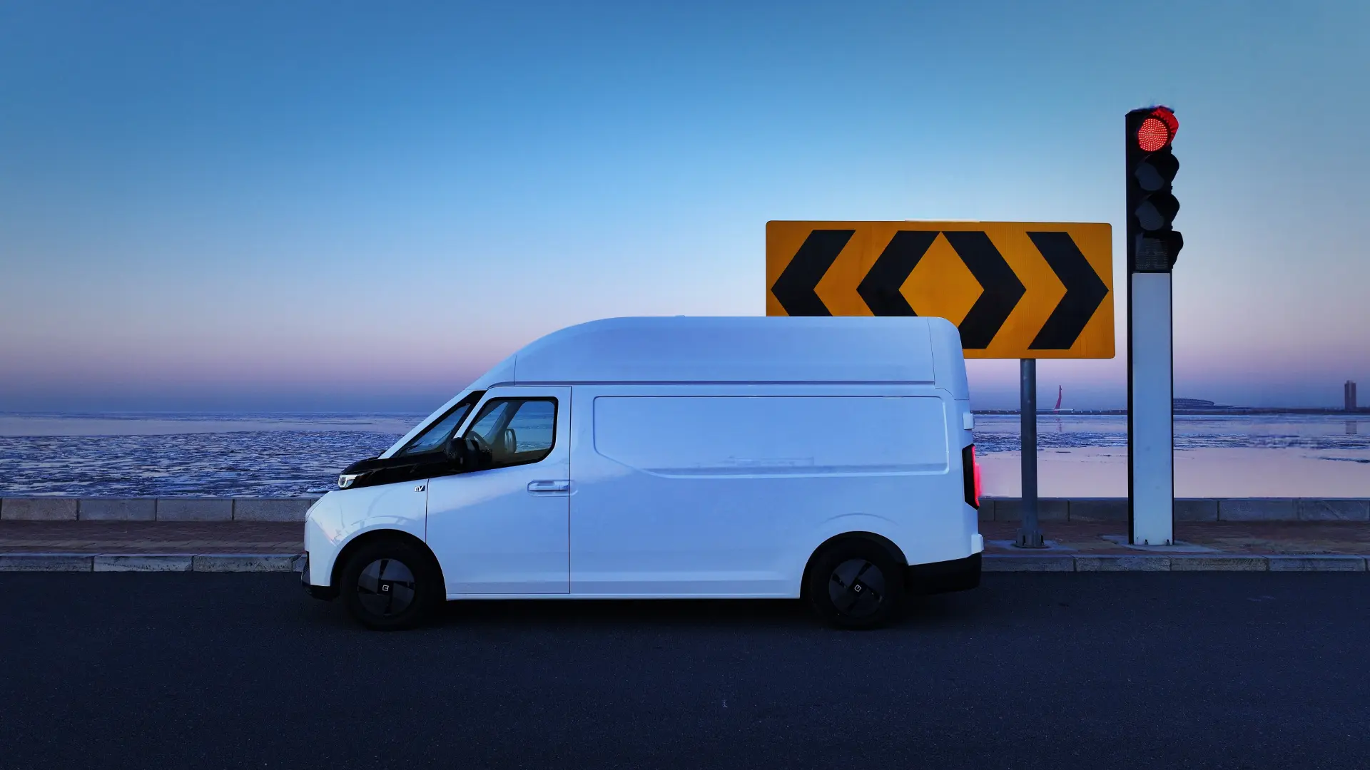 Una camioneta blanca se detuvo en un semáforo en rojo junto a una señal de tráfico con forma de chevron, con una vista de la costa de fondo al anochecer, casi como una corriente automática de serena belleza nocturna.