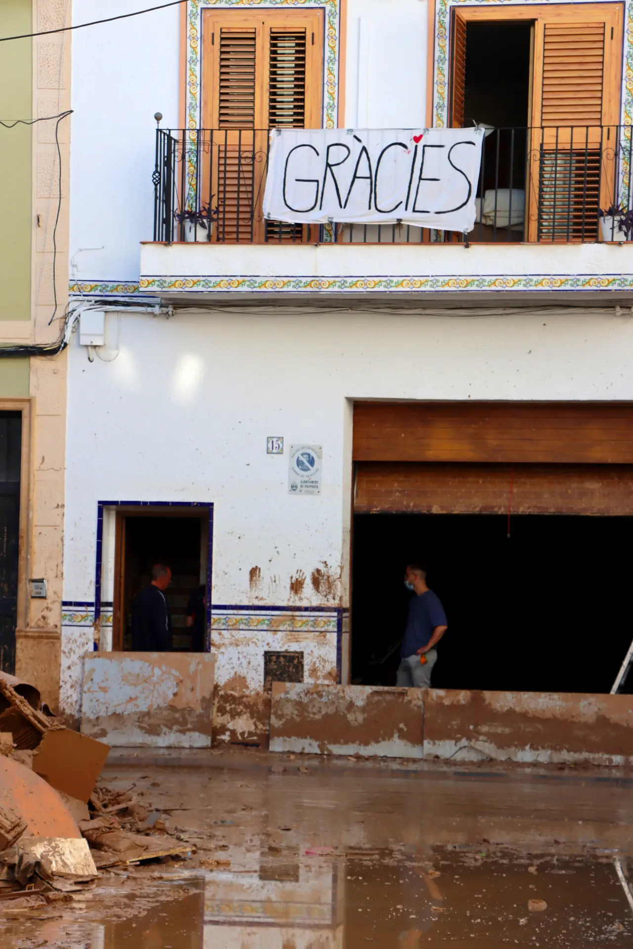 Dos personas limpian una calle inundada, probablemente afectada por un reciente diluvio. Un gran cartel que dice "Gràcies" cuelga de un balcón sobre la entrada embarrada, quizás como agradecimiento al sector del transporte por sus incansables esfuerzos en condiciones tan difíciles.