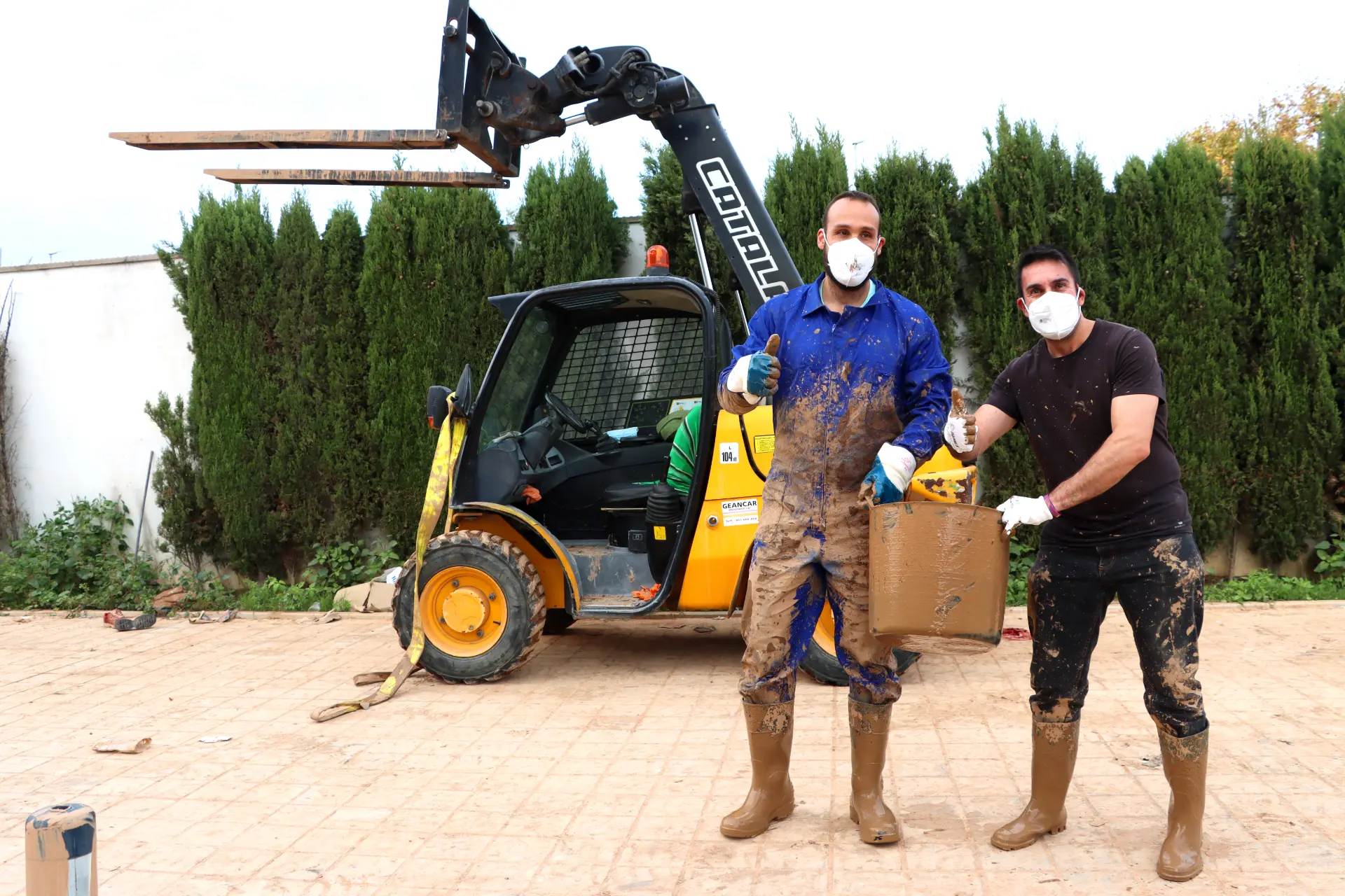 Dos personas con ropa de trabajo embarrada y mascarillas se mantienen de pie orgullosas junto a un cargador que sostiene una caja cubierta de barro y hacen un gesto con el pulgar hacia arriba. Está claro que un día difícil no detendrá su dedicación al sector del transporte, ya que superan los obstáculos con determinación.