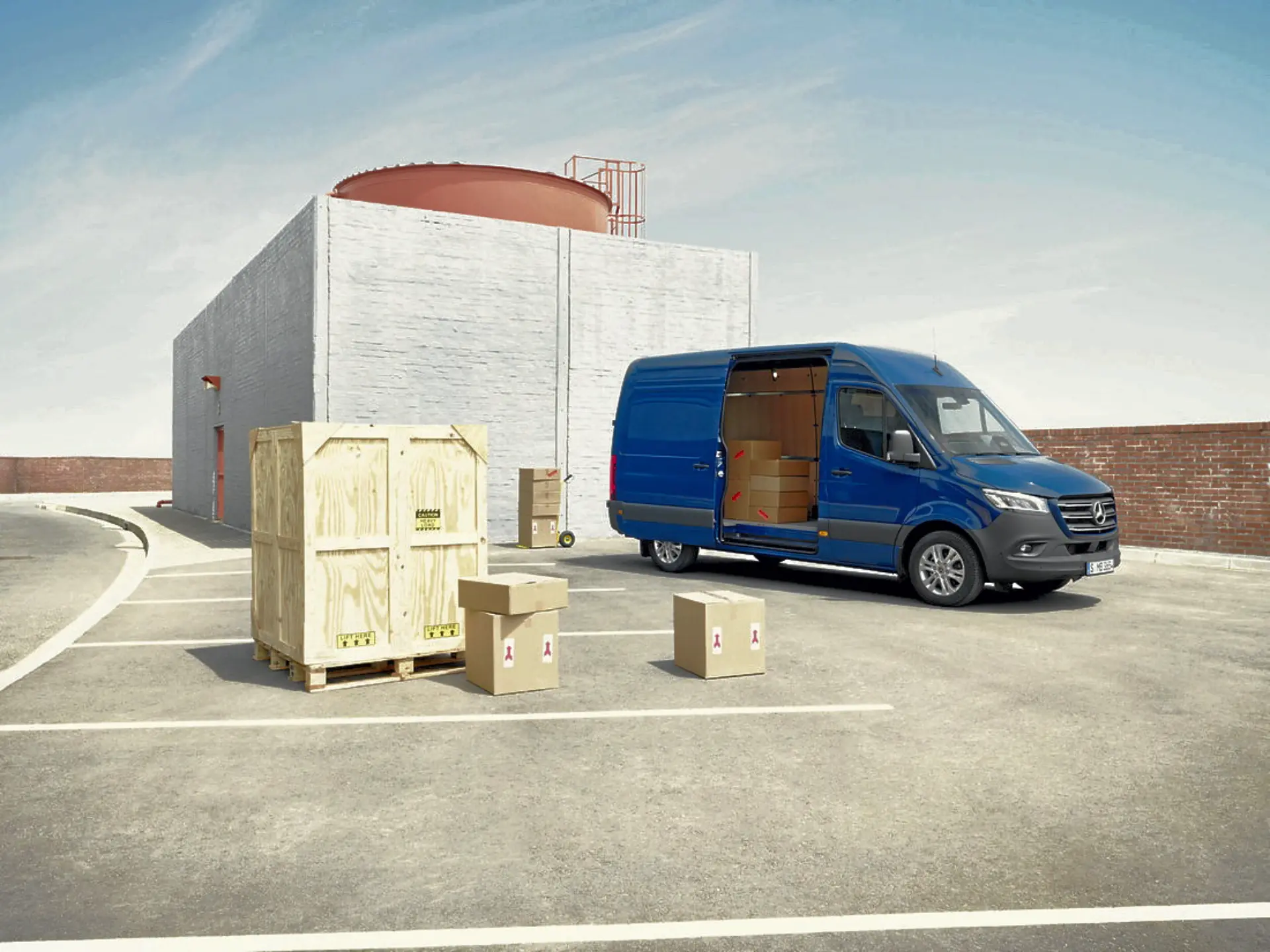 Un Mercedes-Benz Sprinter azul con las puertas abiertas está estacionado junto a un edificio. Cerca de allí se encuentran cajas de madera y de cartón listas para las entregas del día de profesionales dedicados a su oficio.