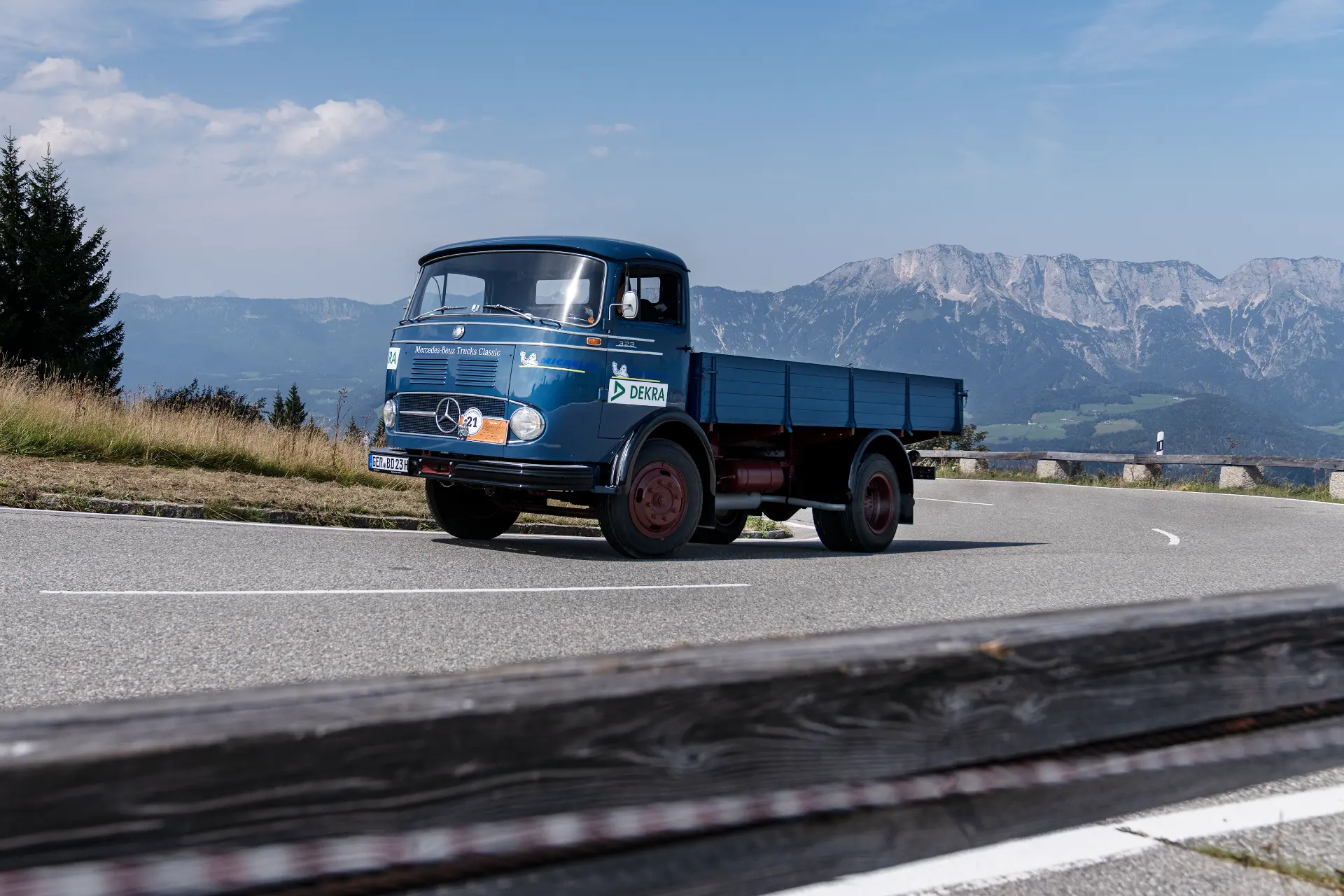 Un camión antiguo azul serpentea por una carretera de montaña, con colinas y un cielo espectacular como telón de fondo. Es como si la naturaleza misma hubiera sido dibujada por un borrador automático, capturando un momento de tranquilidad pura.