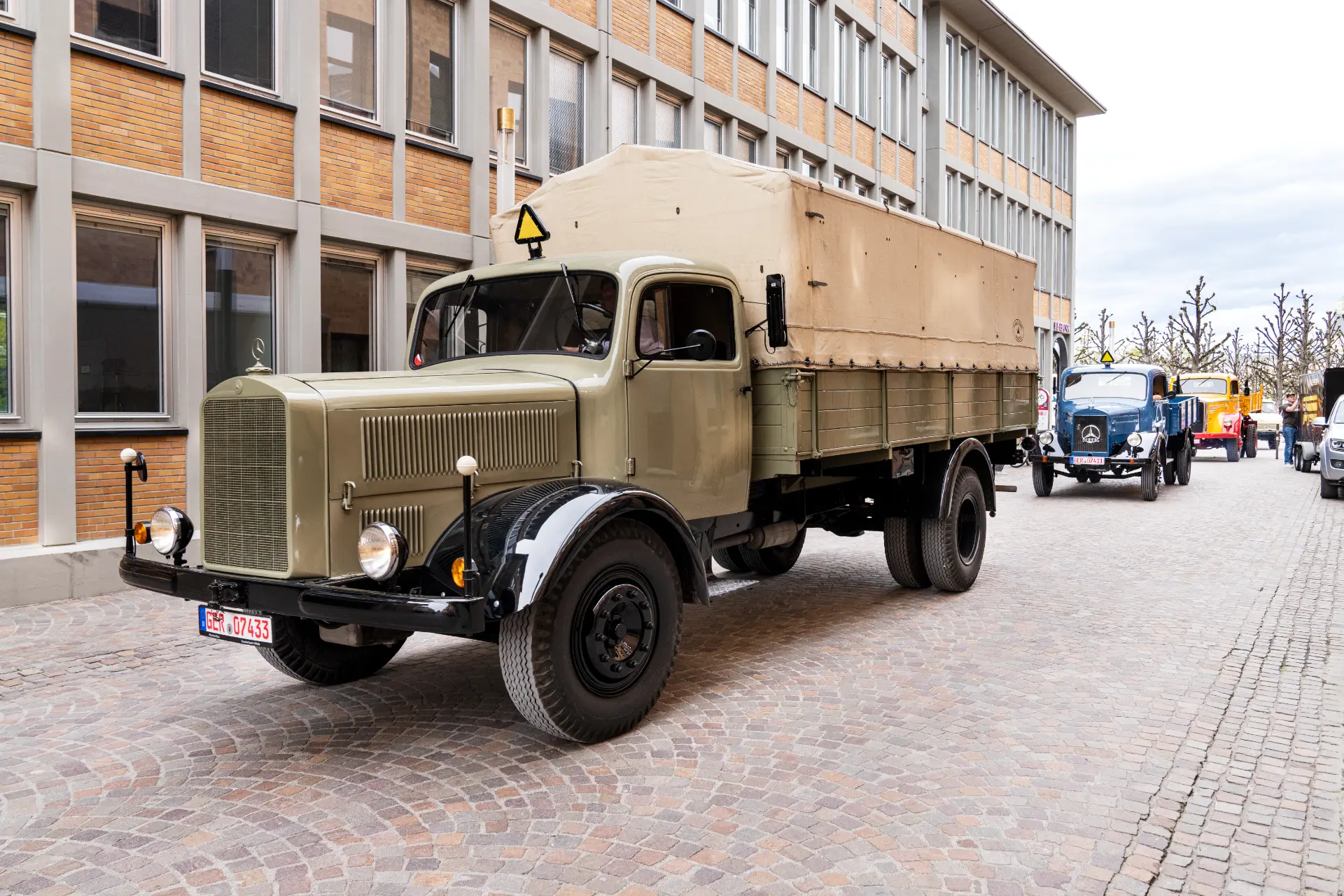 Un camión militar antiguo con un área de carga cubierta de color beige circula por una calle adoquinada, recordando una época en la que no todo era automático.