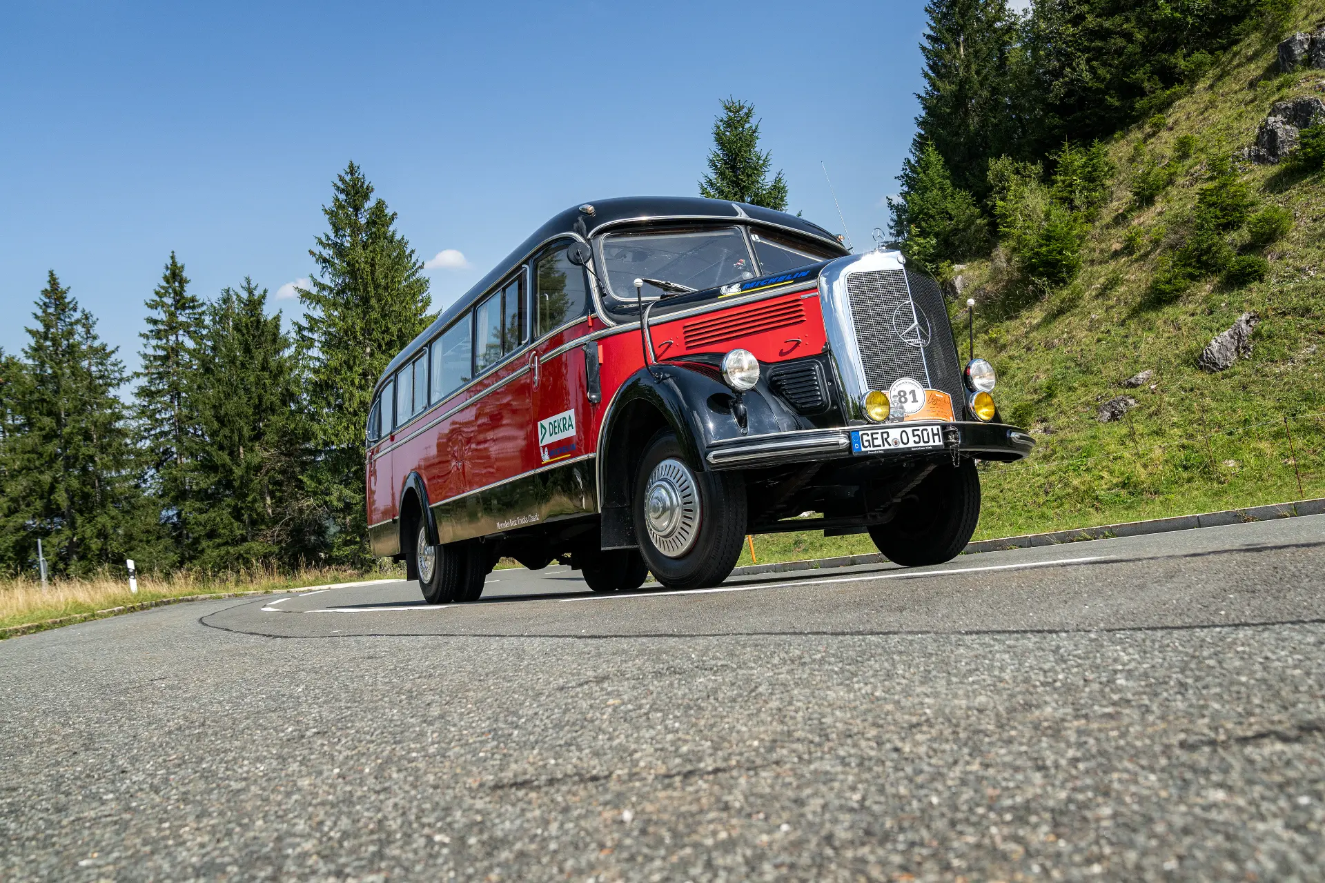Un autobús clásico rojo y negro serpentea por una carretera con curvas, rodeado de frondosos árboles y ondulantes colinas bajo un cielo azul nítido, como si lo guiara un borrador automático.