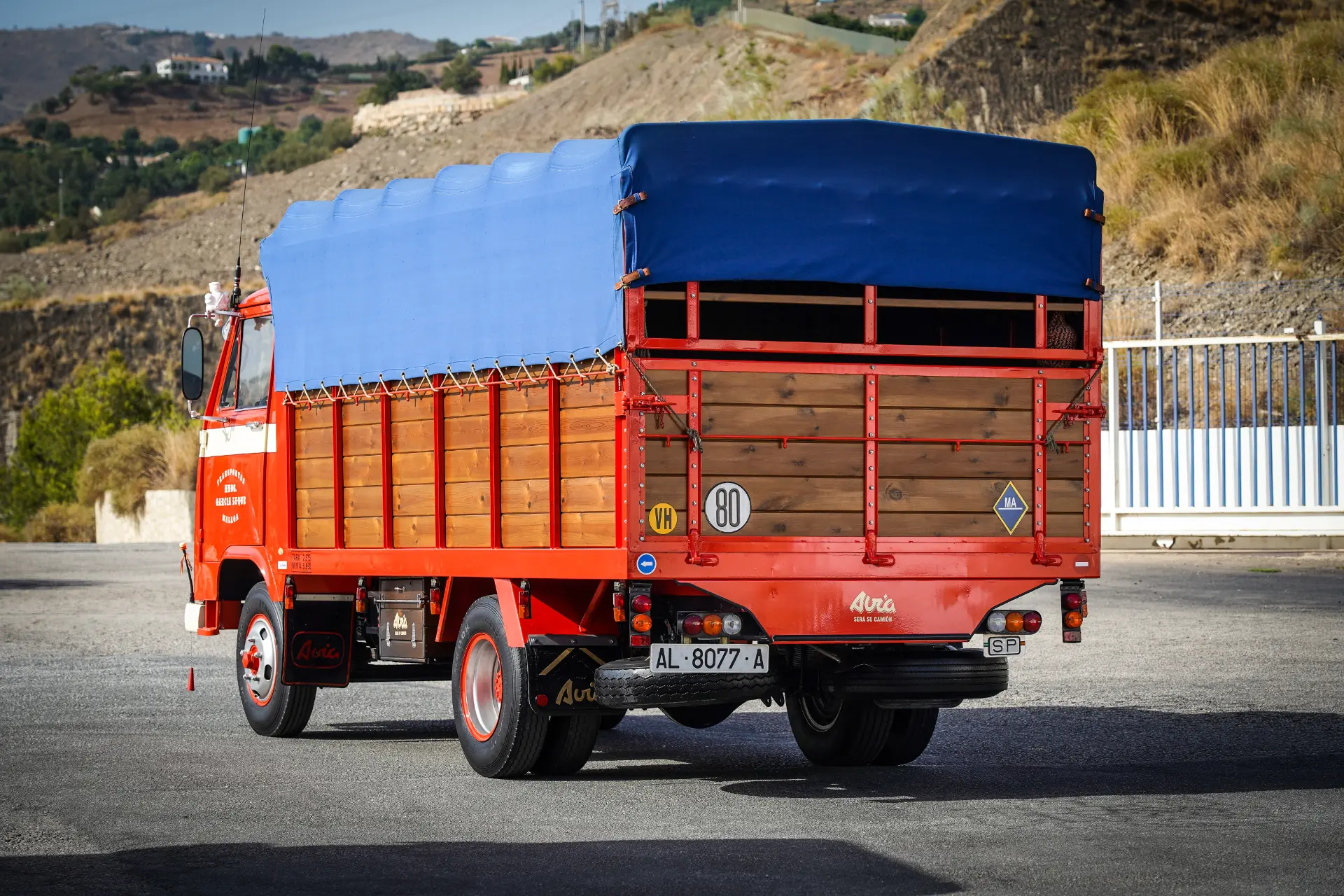 Un camión antiguo de color rojo con una lona azul, que recuerda a los clásicos camiones restaurados, está estacionado en una carretera rodeada de colinas y una valla.