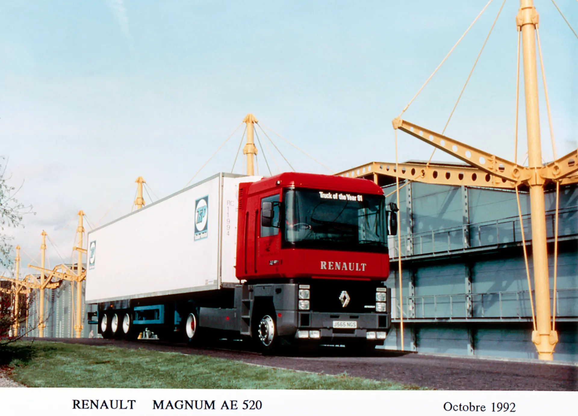 Camión Renault Magnum AE 520 rojo con remolque blanco cerca de un moderno almacén amarillo de acero y cristal. Esta vista nos remonta a octubre de 1992, mucho antes de la era de las innovaciones de Renault Trucks, como los camiones eléctricos Renault E-Tech.