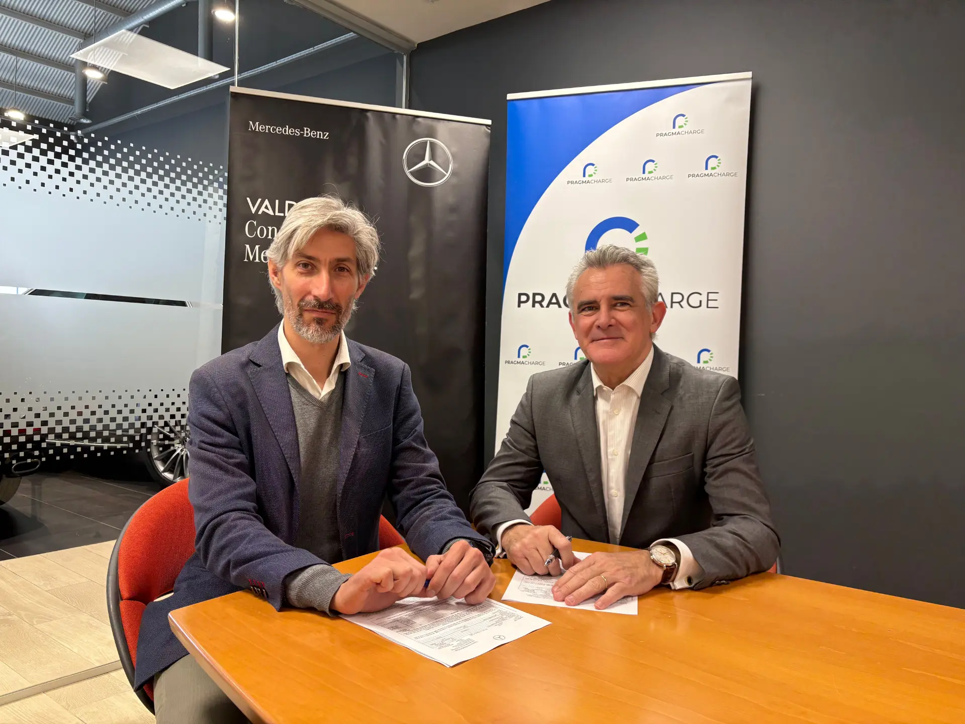 Dos hombres de traje se sientan en una mesa con documentos, con pancartas de Mercedes-Benz y PRA Group en el fondo, discutiendo el futuro de los camiones eléctricos en España y el impacto potencial del Mercedes-Benz eActros 600 en la construcción de una flota sostenible PragmaCharge.