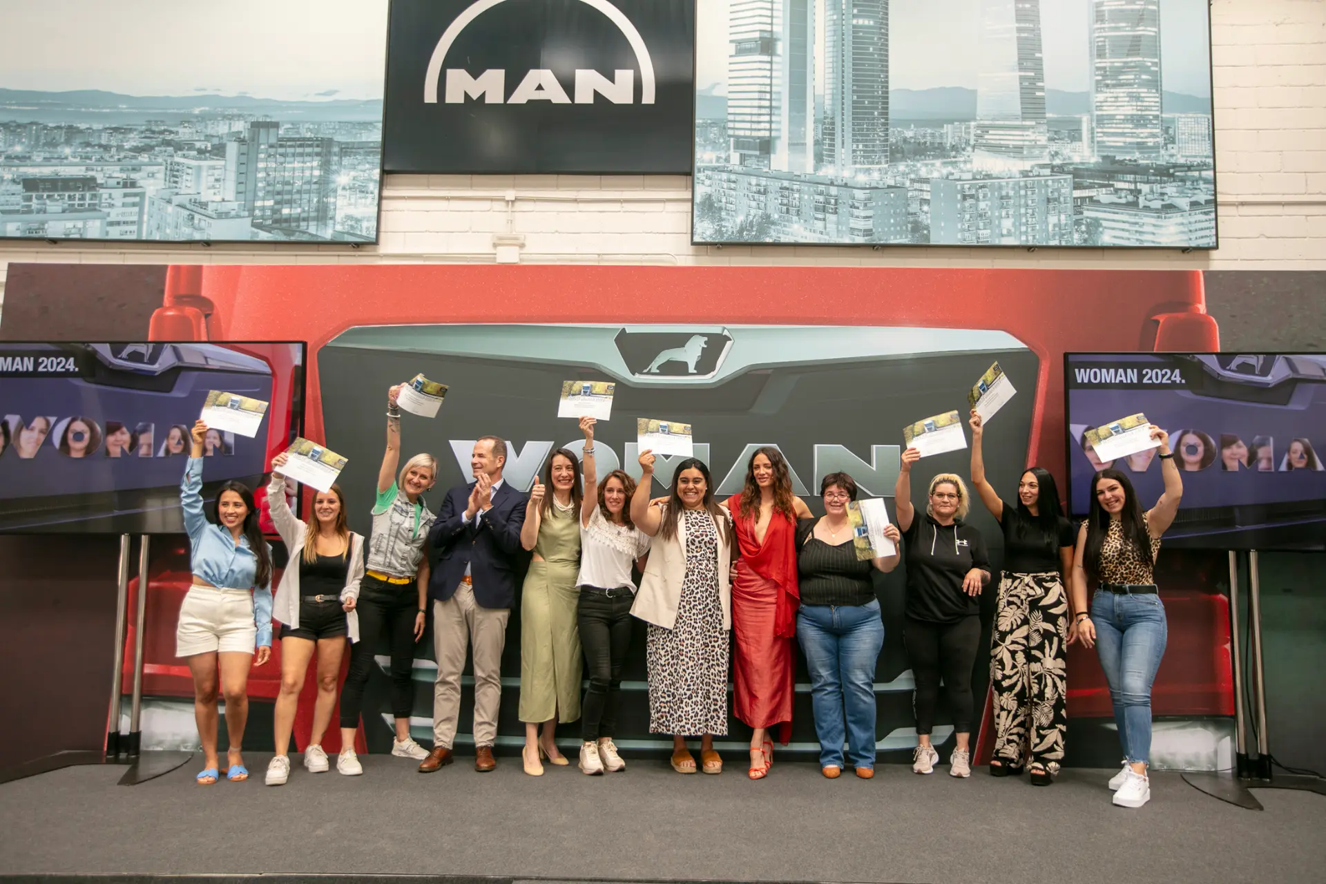 Un grupo de personas sosteniendo orgullosamente sus certificados, sonriendo y posando en el escenario frente a un cartel de MAN; representan el éxito colectivo de una innovación de borrador automático.
