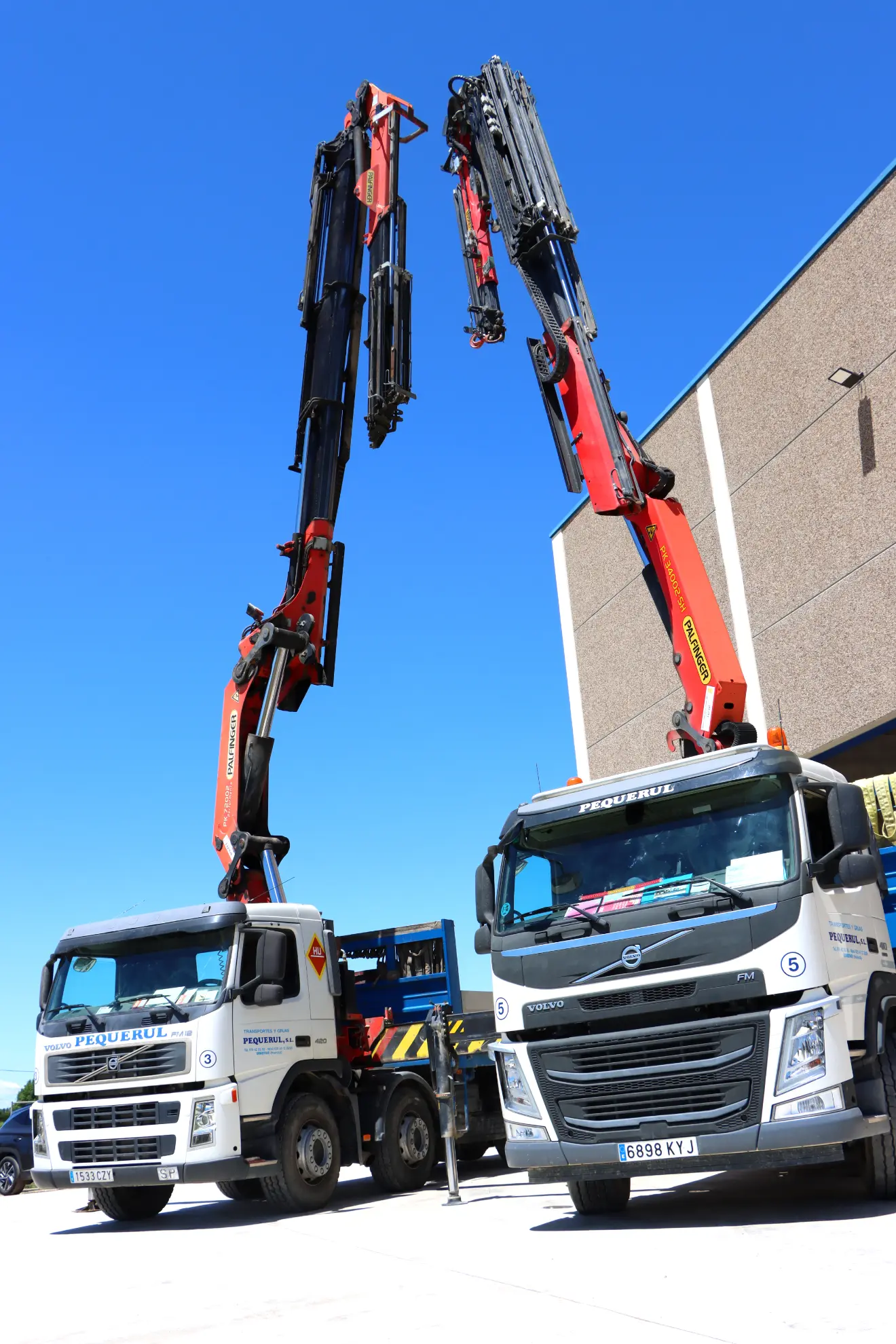 Dos grandes camiones con grúas extendidas, parte de una operación de borrador automático, se enfrentan afuera de un edificio bajo un cielo azul claro.