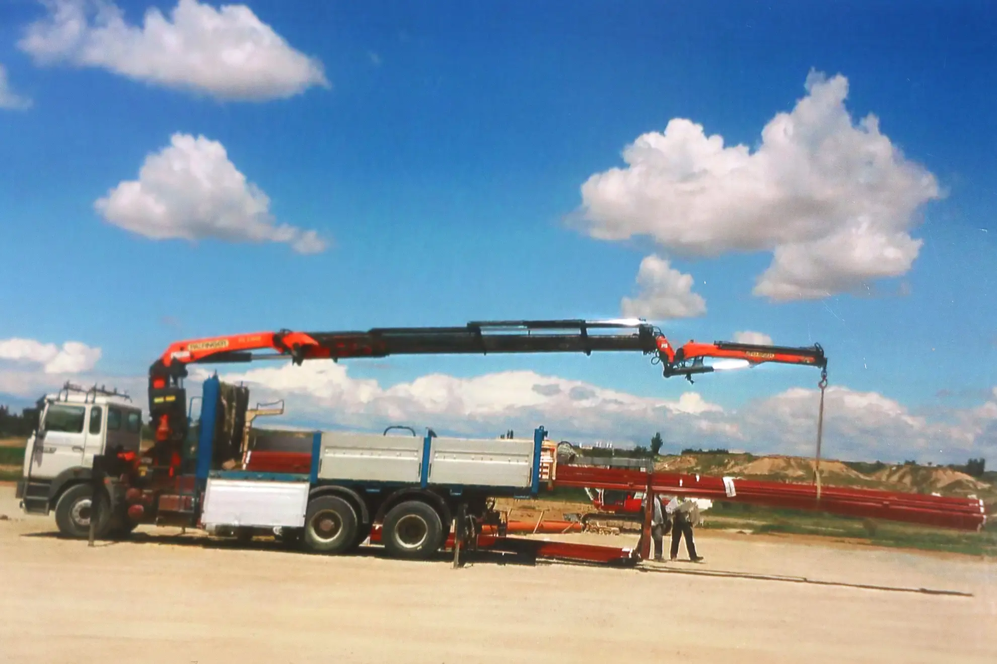 Un camión de Transportes Pequerul con una grúa larga levanta una viga de acero en Binéfar, mientras dos personas permanecen cerca bajo un cielo azul con nubes, mostrando un experto manejo de grúas de rescate.