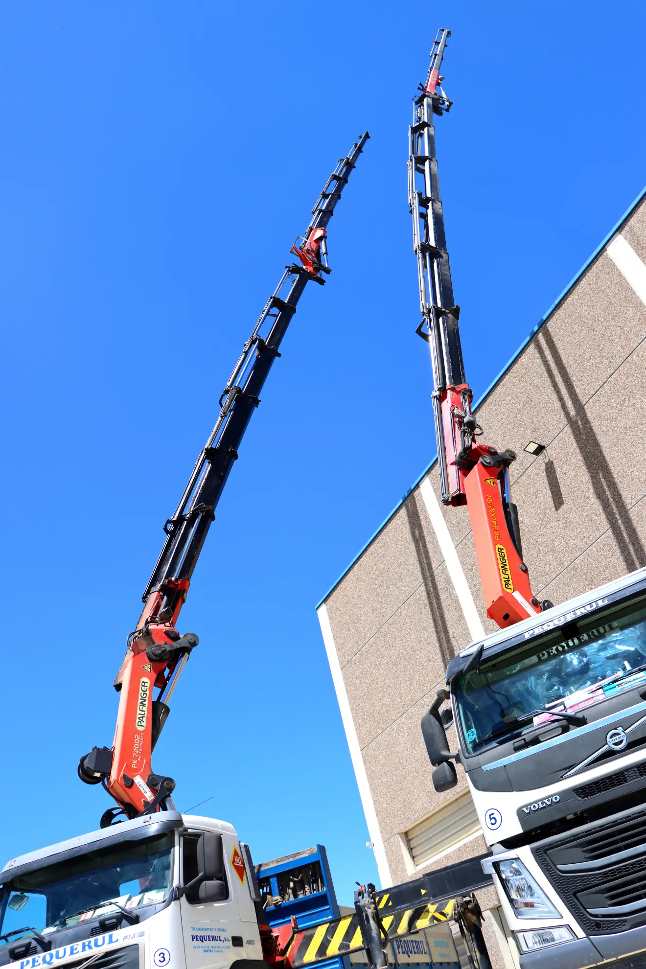 Dos camiones con grúas de rescate extendidas de Transportes Pequerul se elevan contra un cielo azul claro cerca de un edificio en Huesca.