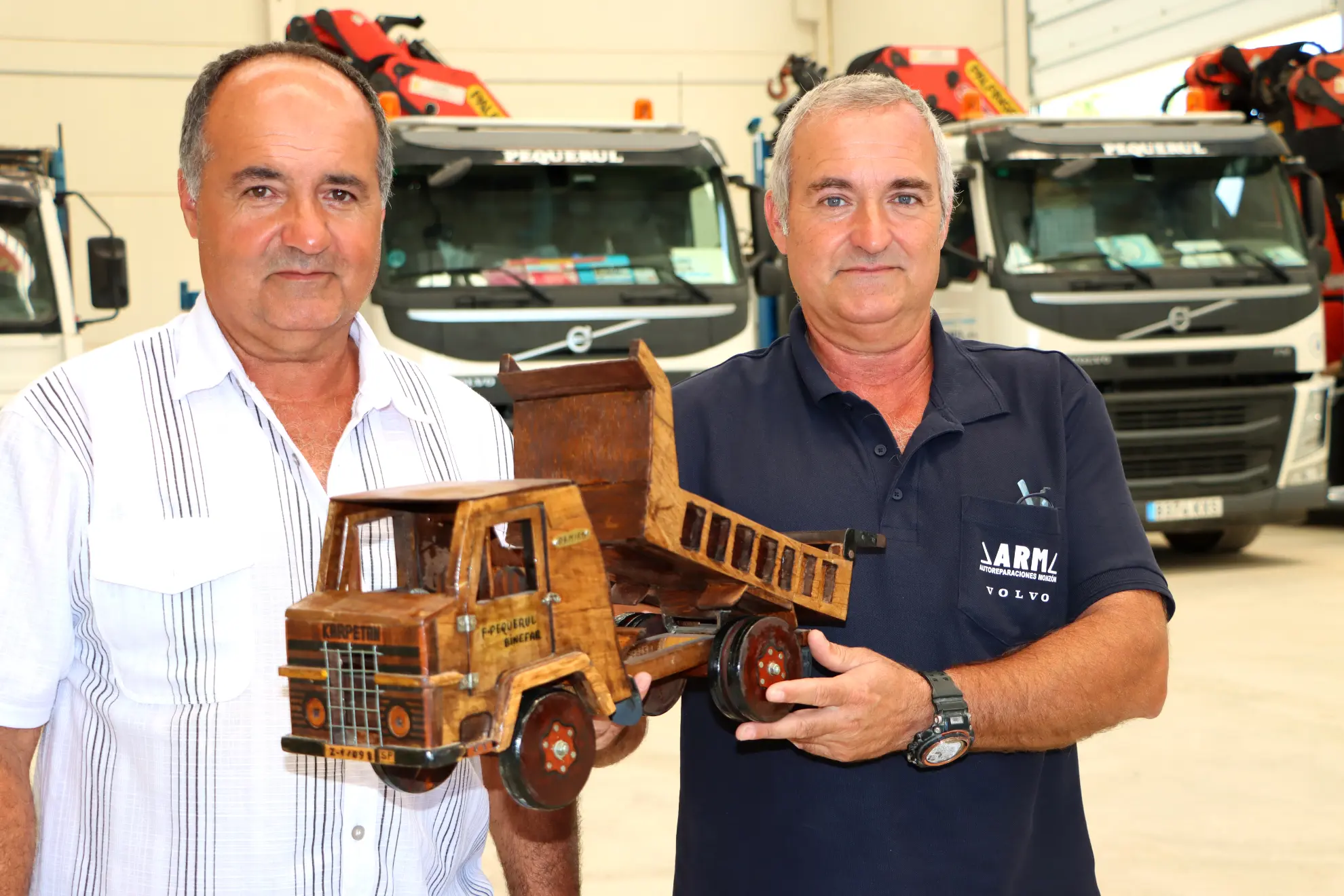 Dos hombres se encuentran en un interior frente a camiones, sosteniendo con orgullo un modelo detallado de madera de un camión volquete, un ejemplo impresionante de la artesanía del Borrador automático.