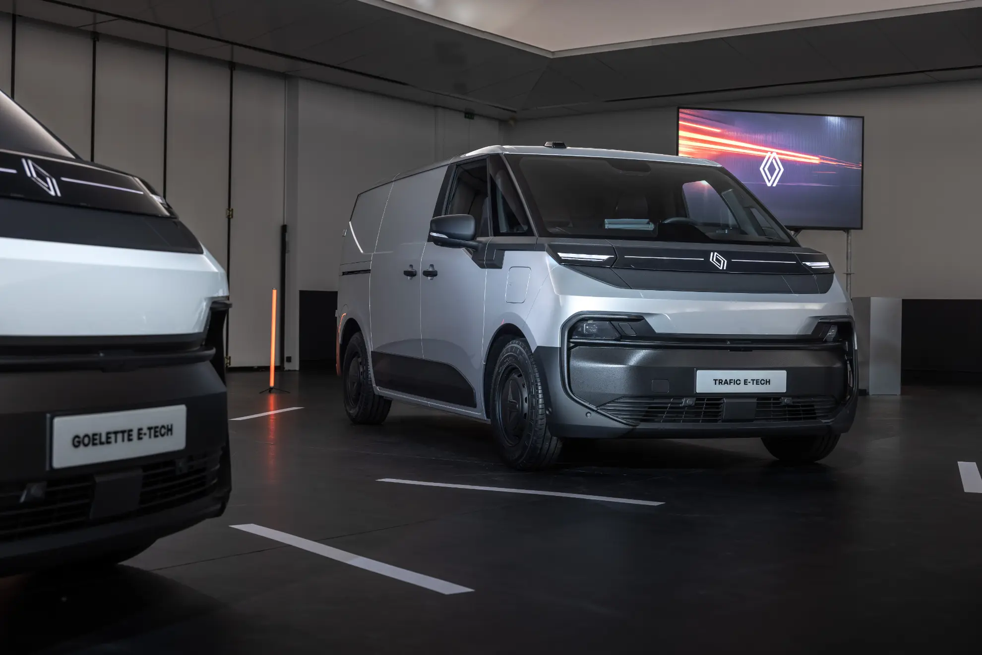 Furgoneta Renault Trafic E-Tech color plata expuesta en el interior con otro vehículo parcialmente visible a la izquierda, que incorpora un sistema de borrador automático para mayor comodidad.