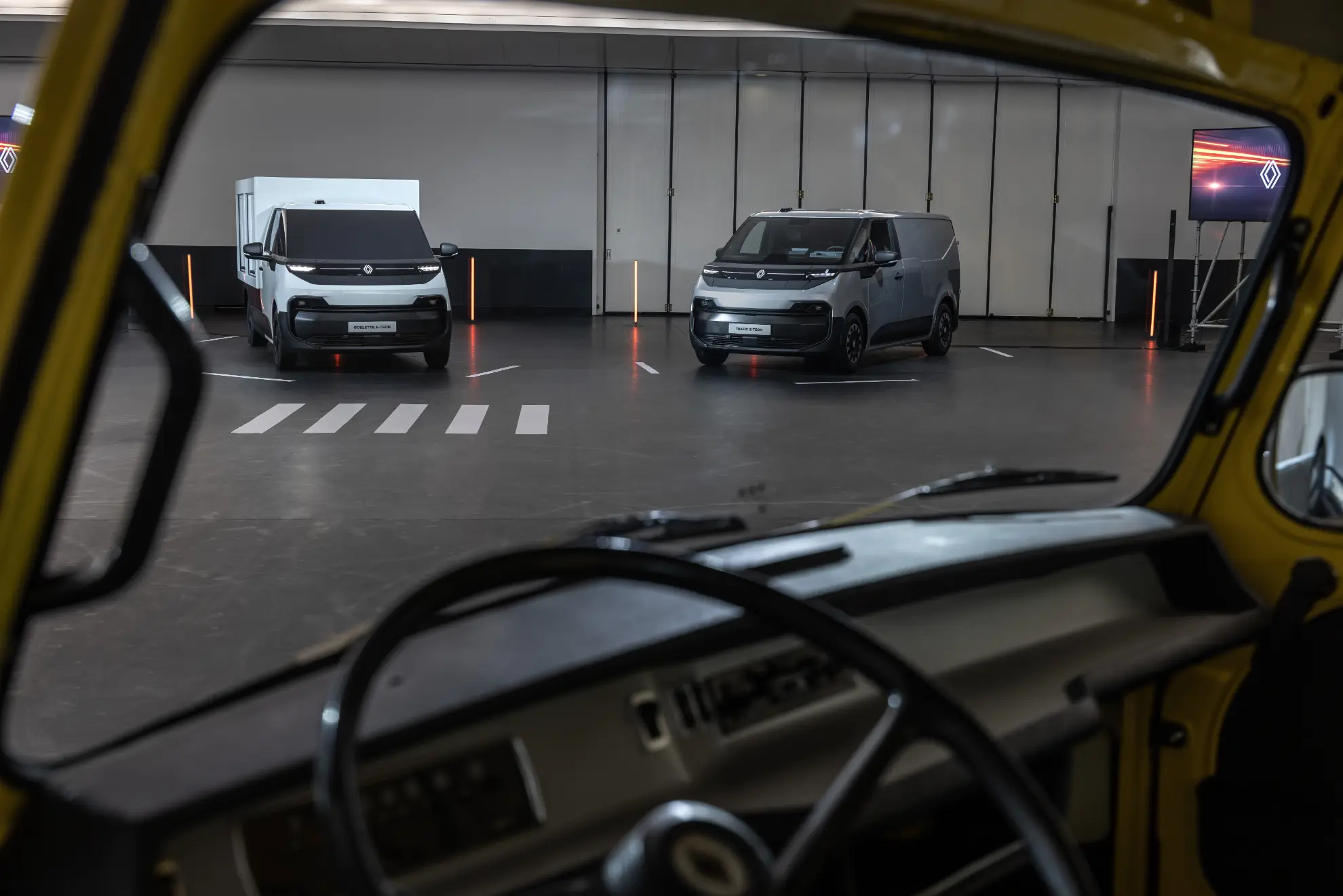 Vista desde el interior de una furgoneta clásica, mirando hacia dos furgonetas Volkswagen modernas en un gran espacio interior, capturado como un borrador automático.