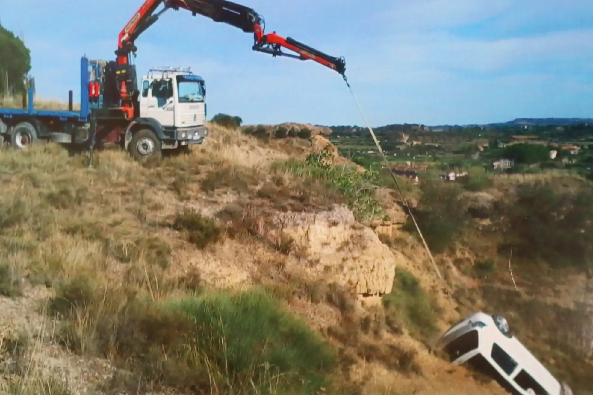 Una grúa de Transportes Pequerul levanta un coche desde una ladera empinada con un cable en el paisaje rural cerca de Binéfar, mostrando los servicios expertos de las grúas de rescate.