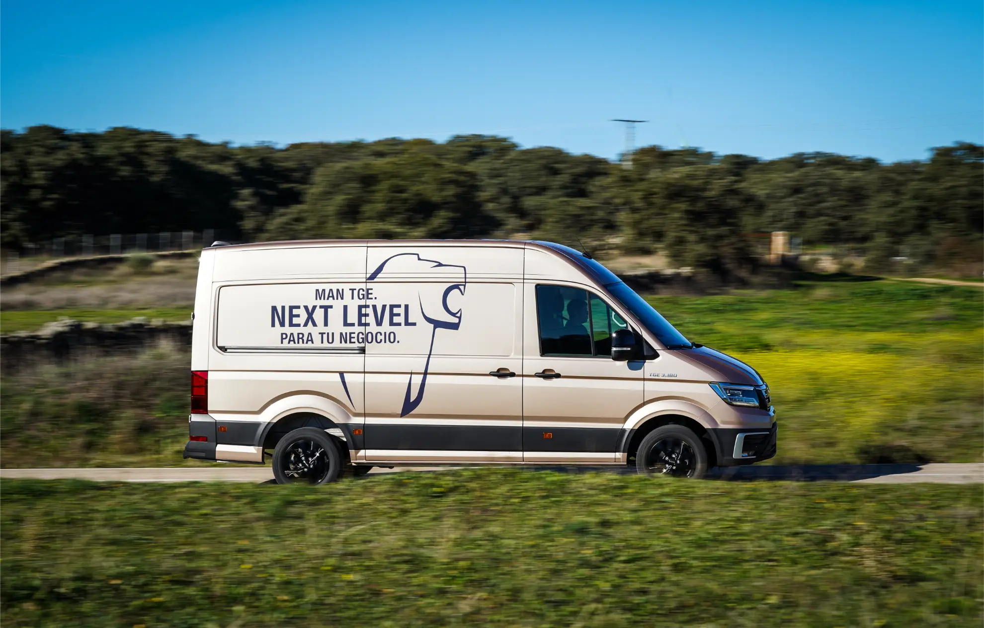 Una furgoneta de reparto MAN TGE 3.180 de color beige con la leyenda "NEXT LEVEL PARA TU NEGOCIO" en el lateral circula por un camino rural junto a la hierba verde.